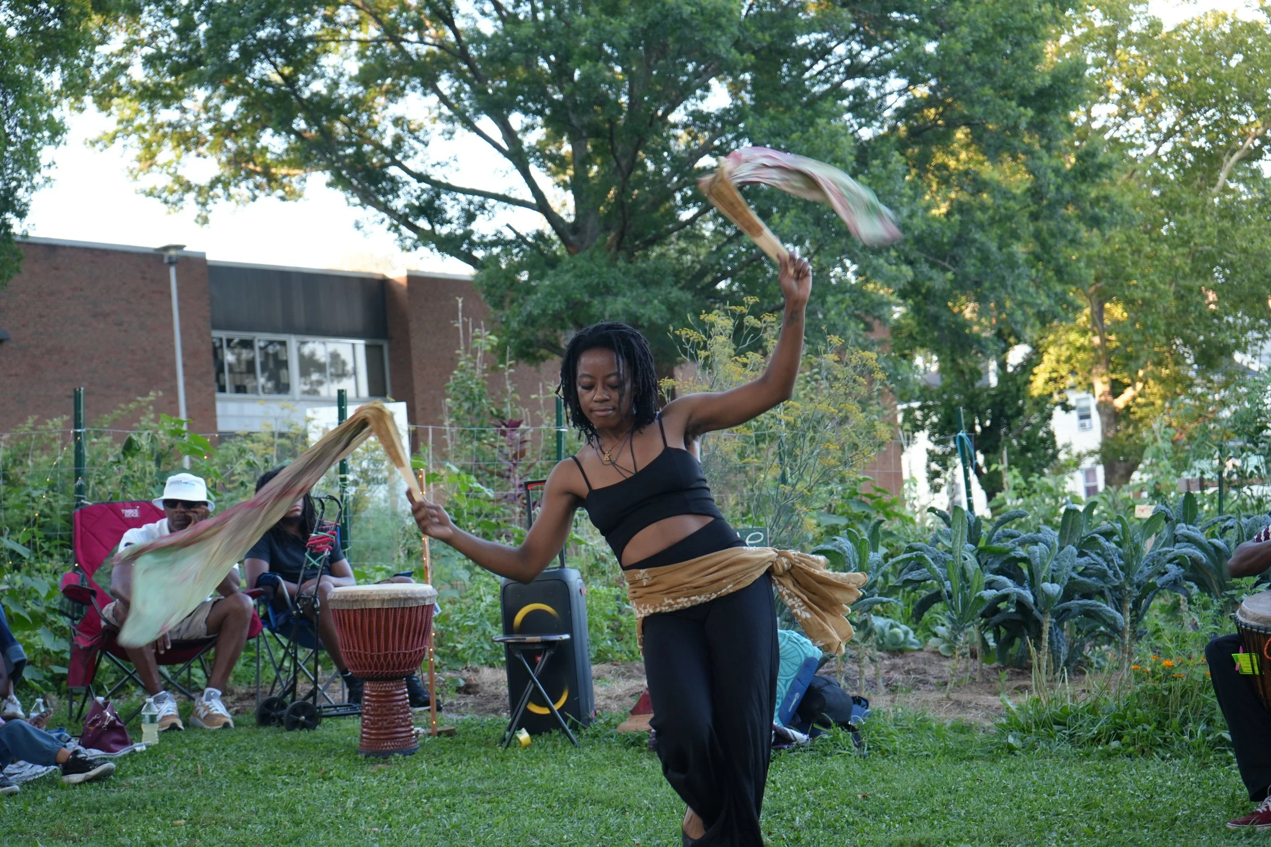 A woman dancing outdoors in a garden, holding fabric in each hand, with music instruments and people seated nearby, surrounded by trees and plants.