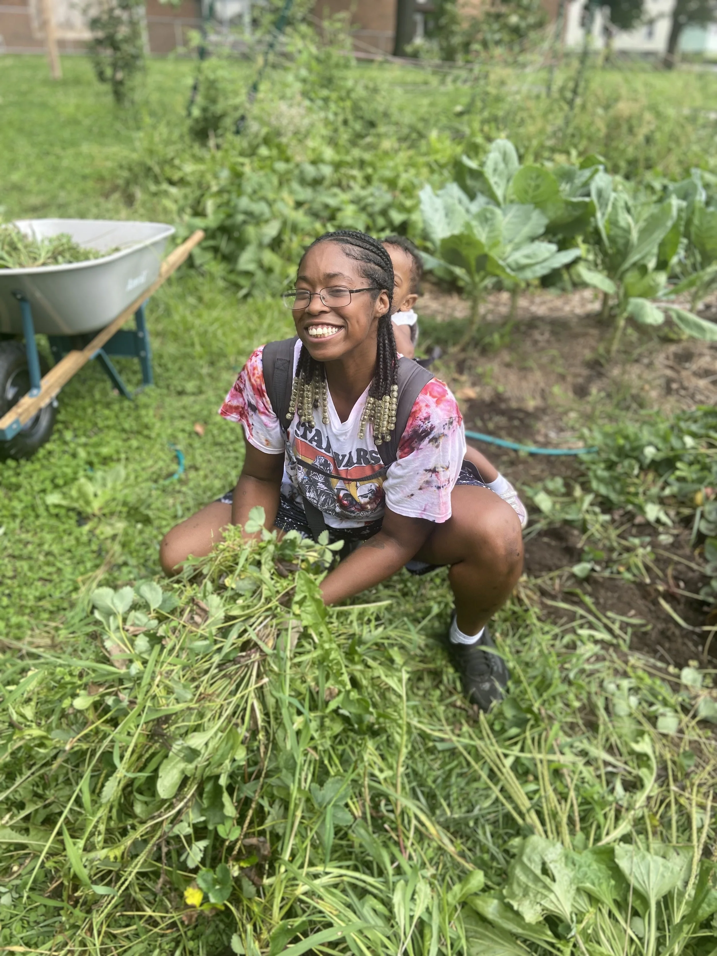 A young woman with braids and glasses squats in a garden, smiling and holding a bunch of green weeds or plants she has pulled out of the ground, with another person partially hidden behind her.