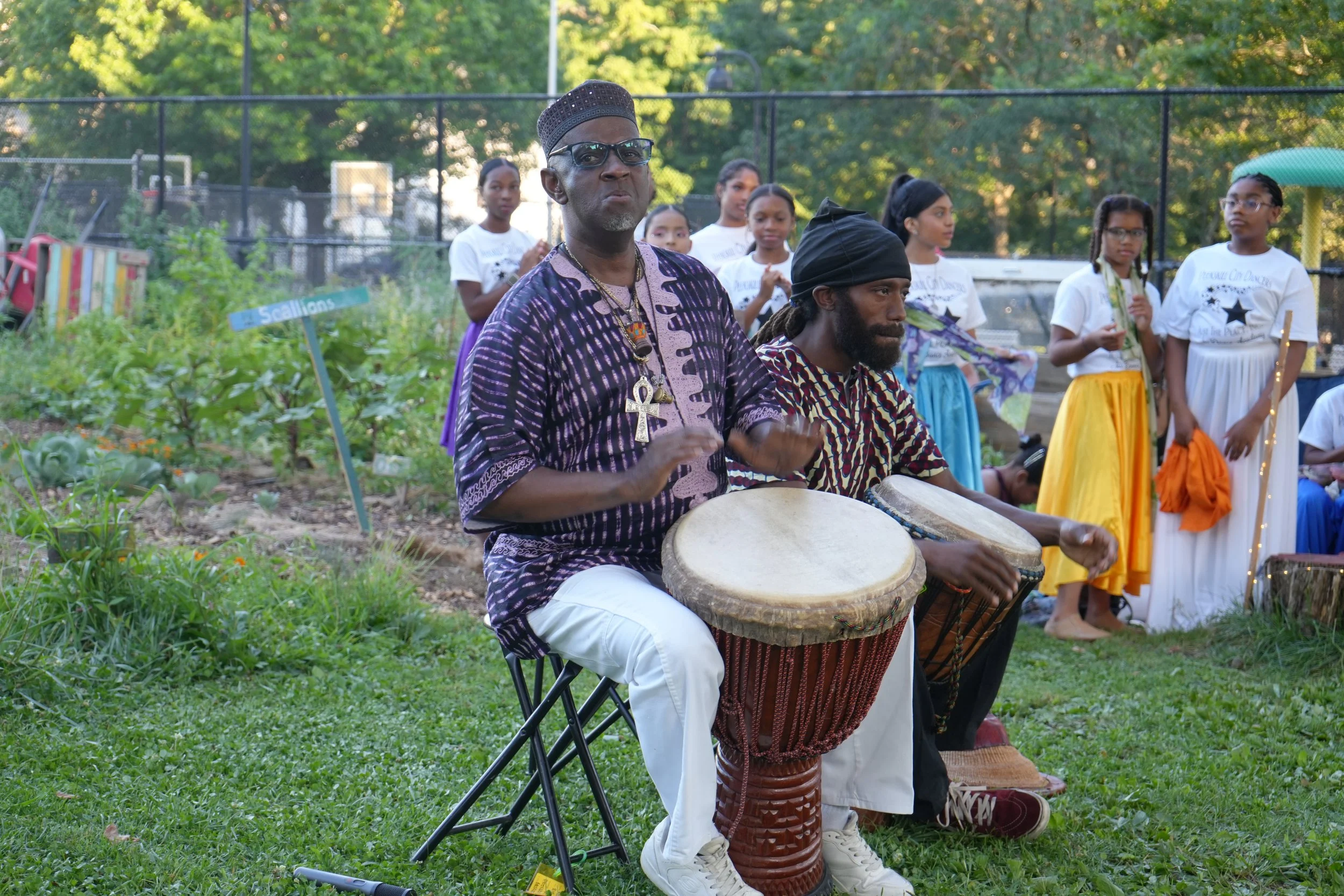 A man wearing sunglasses and a patterned traditional African shirt playing drums, with another man in a similar attire sitting next to him, and a group of children in colorful clothing standing behind them, outdoors in a park with a fence and trees i