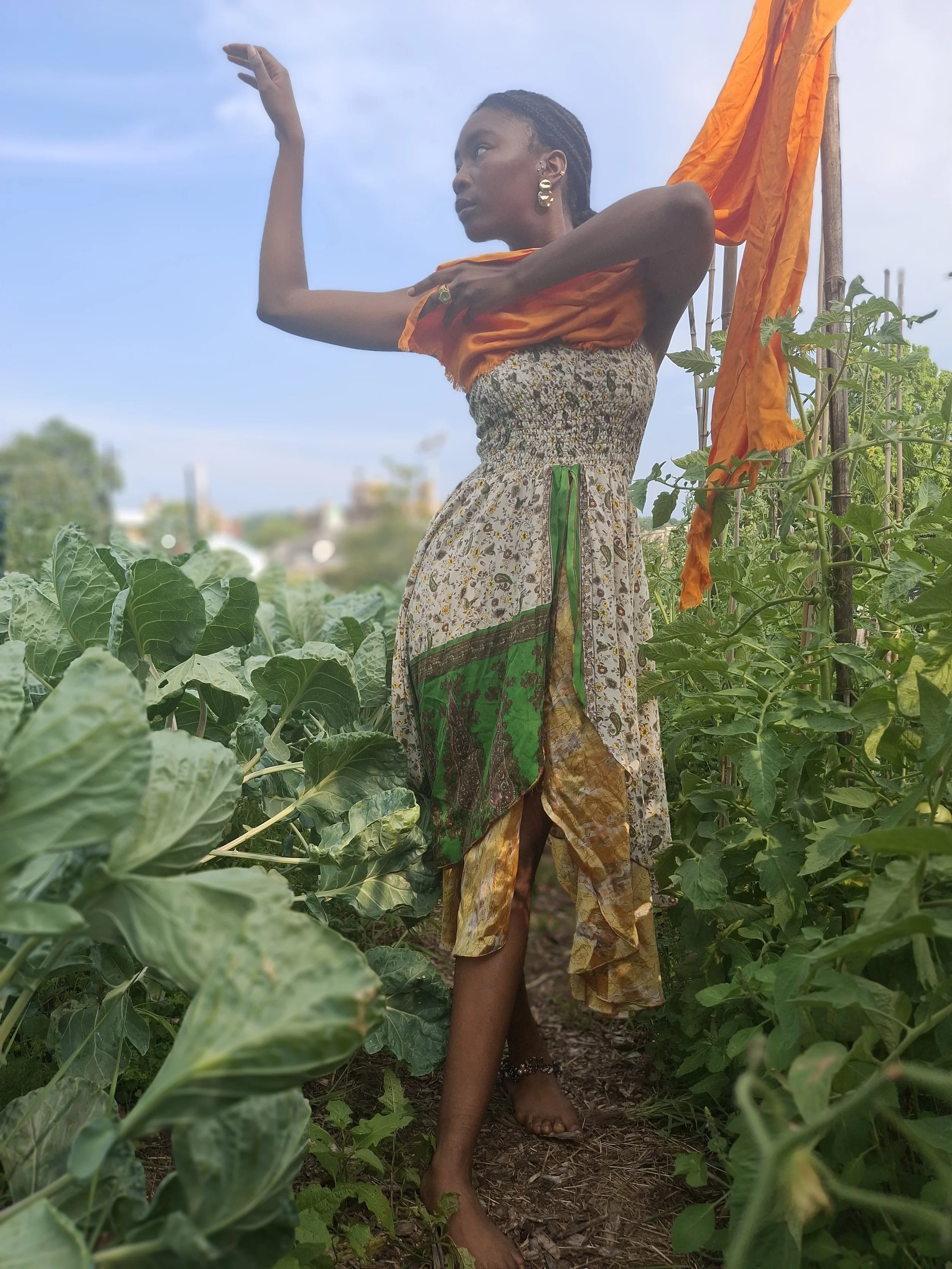 A woman standing barefoot in a garden surrounded by green plants, wearing a colorful dress with orange, green, and earth tones, and striking a dramatic pose with her right arm raised and her left arm crossed over her chest, with a cloudy sky overhead.