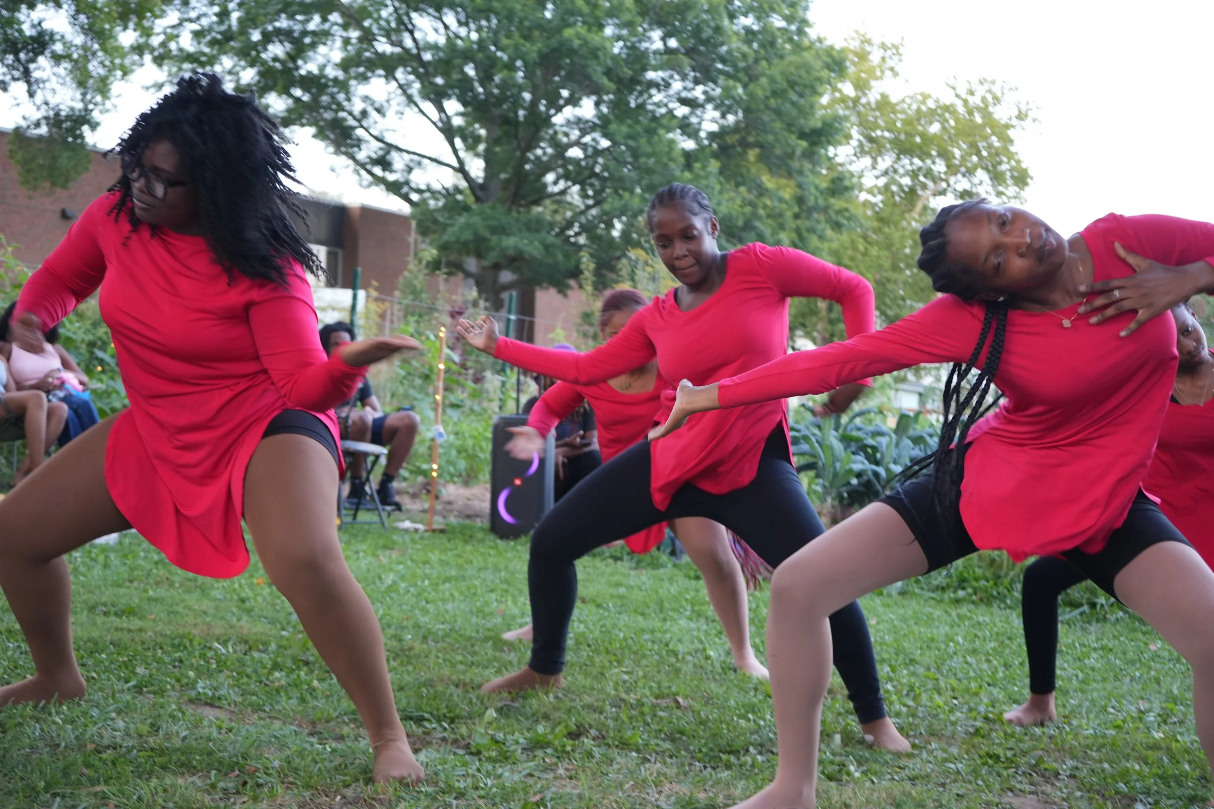 Women in red tops and black bottoms performing a dance outdoors on a grassy area with trees and an audience seated in the background.