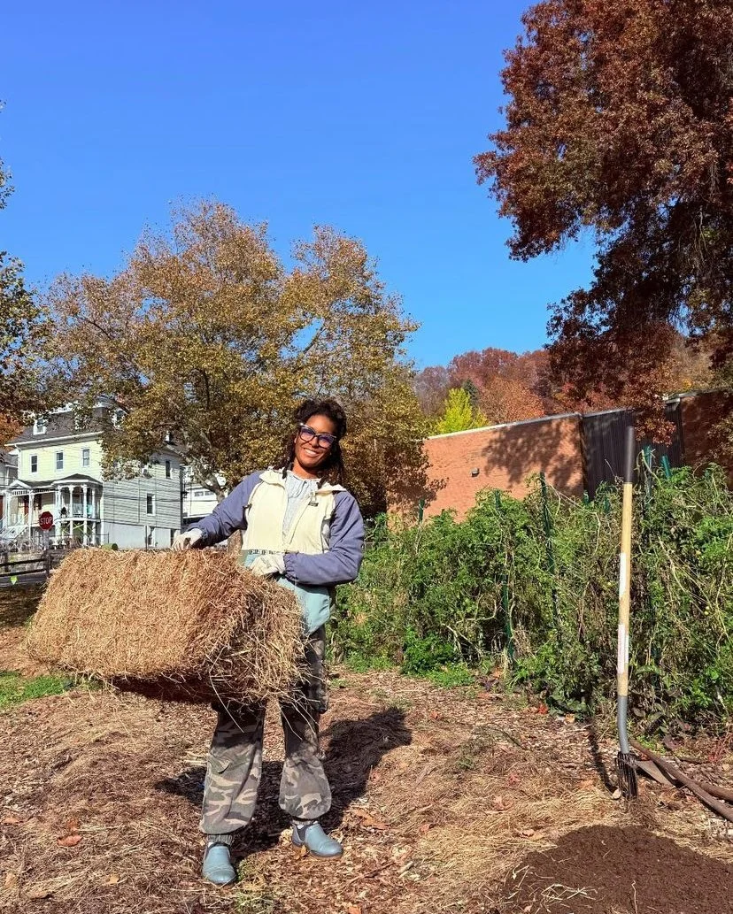 A woman smiling and holding a large hay bale in a garden on a sunny fall day with colorful trees and houses in the background.