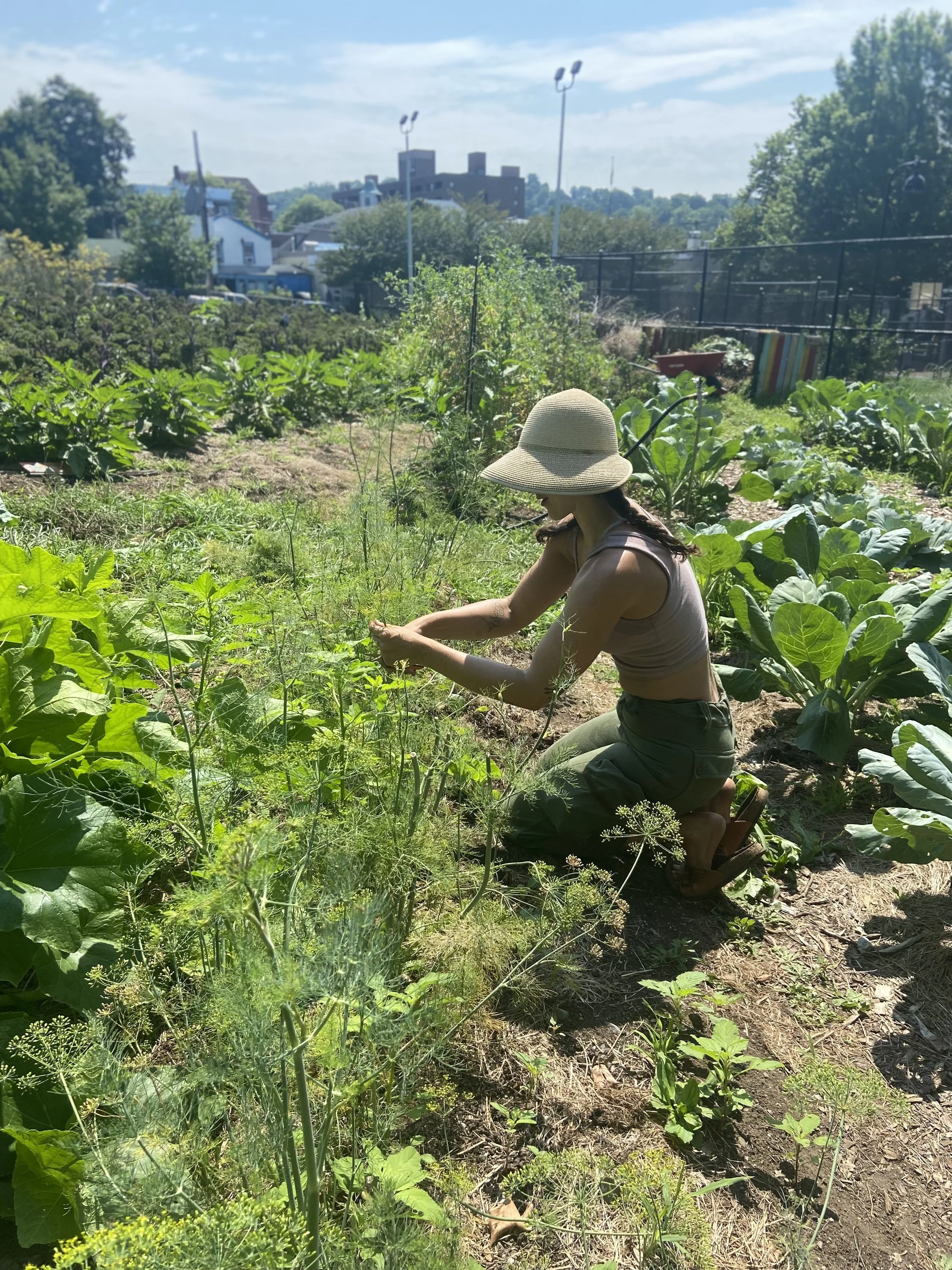 A woman wearing a wide-brimmed hat, tank top, and green pants kneeling in a garden, tending to plants on a sunny day.