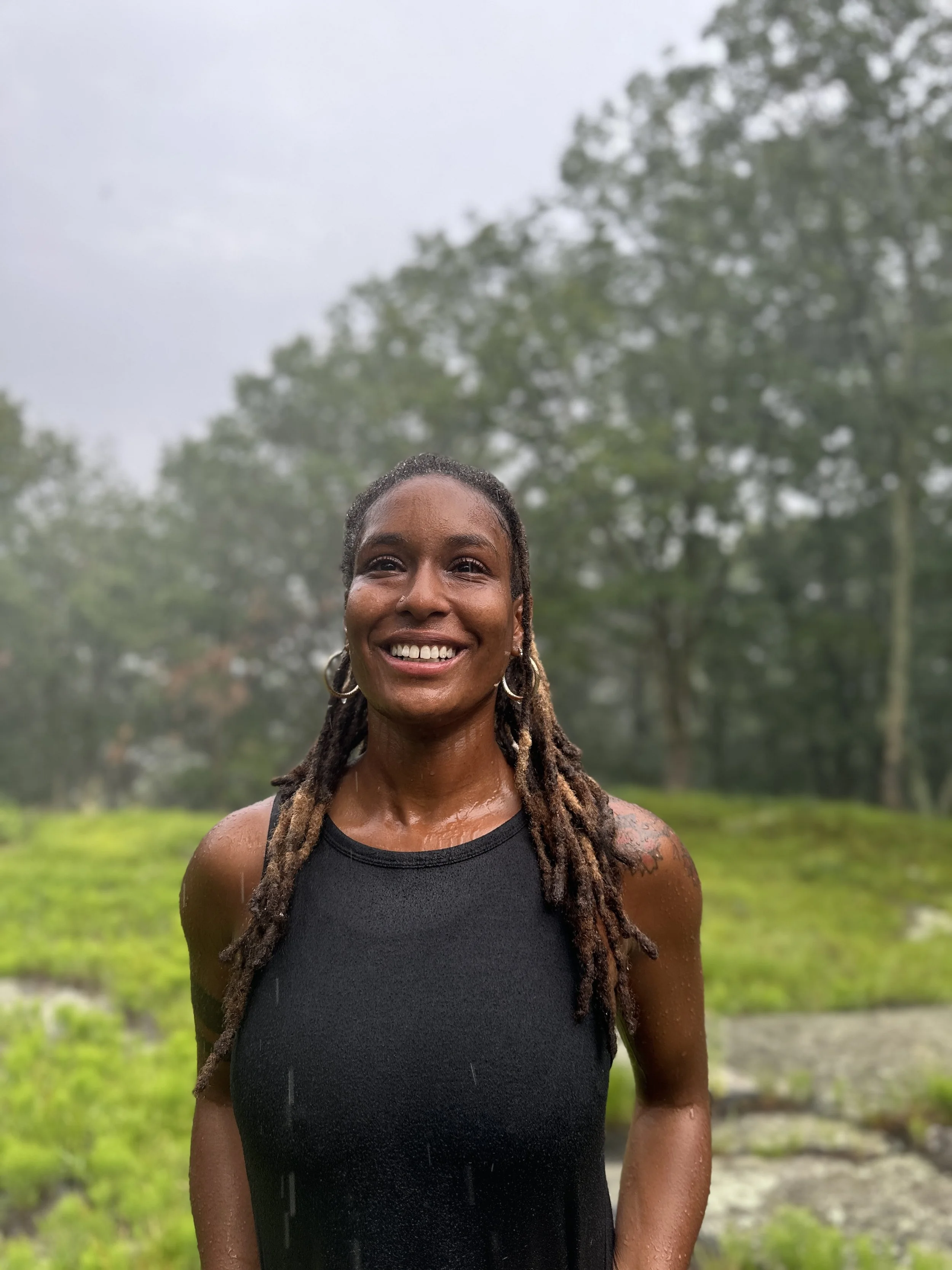 A woman with dreadlocks smiling outdoors in the rain, wearing a black sleeveless top, with trees and greenery in the background.