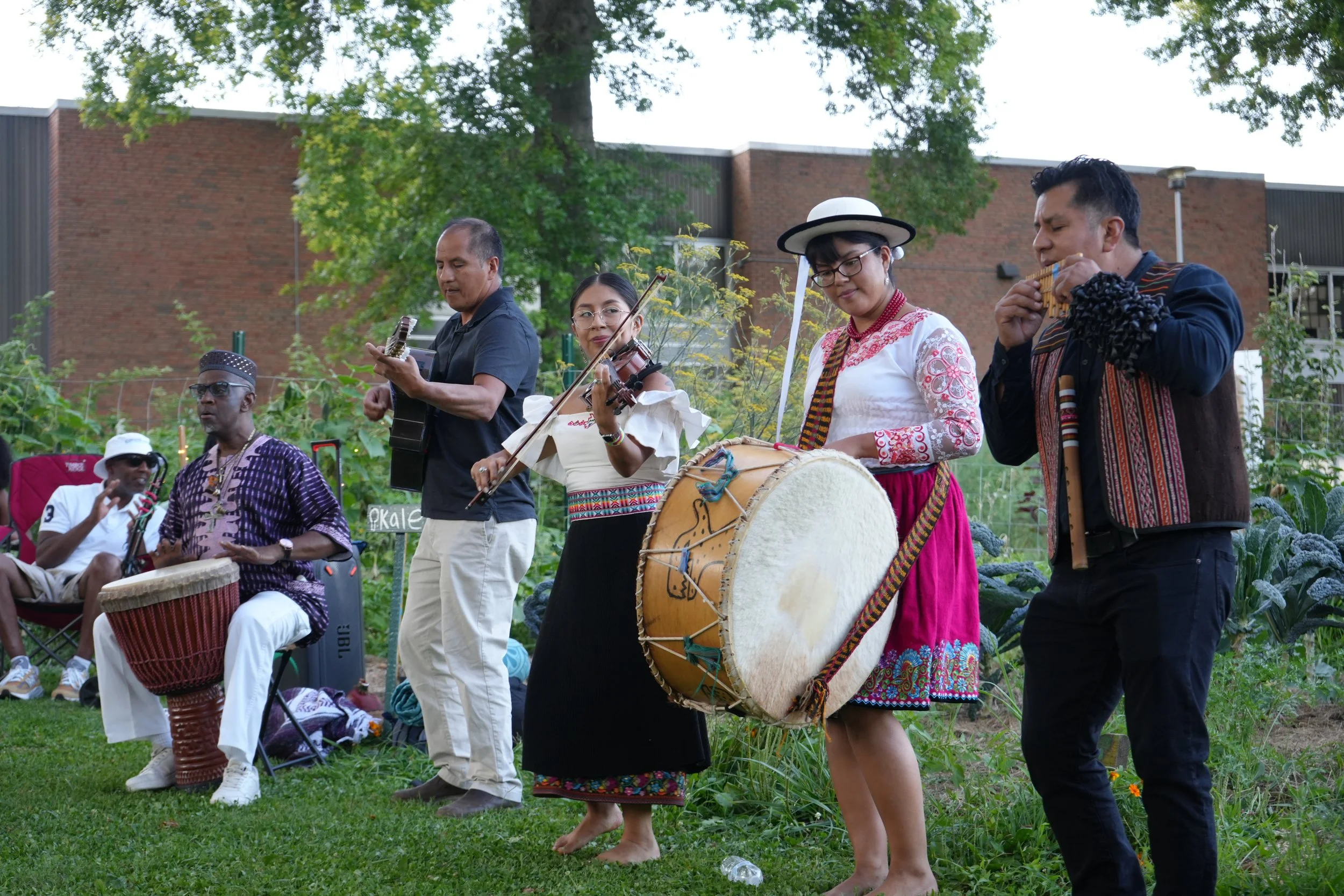 Group of musicians playing instruments outdoors in a garden, with a brick building and trees in the background.