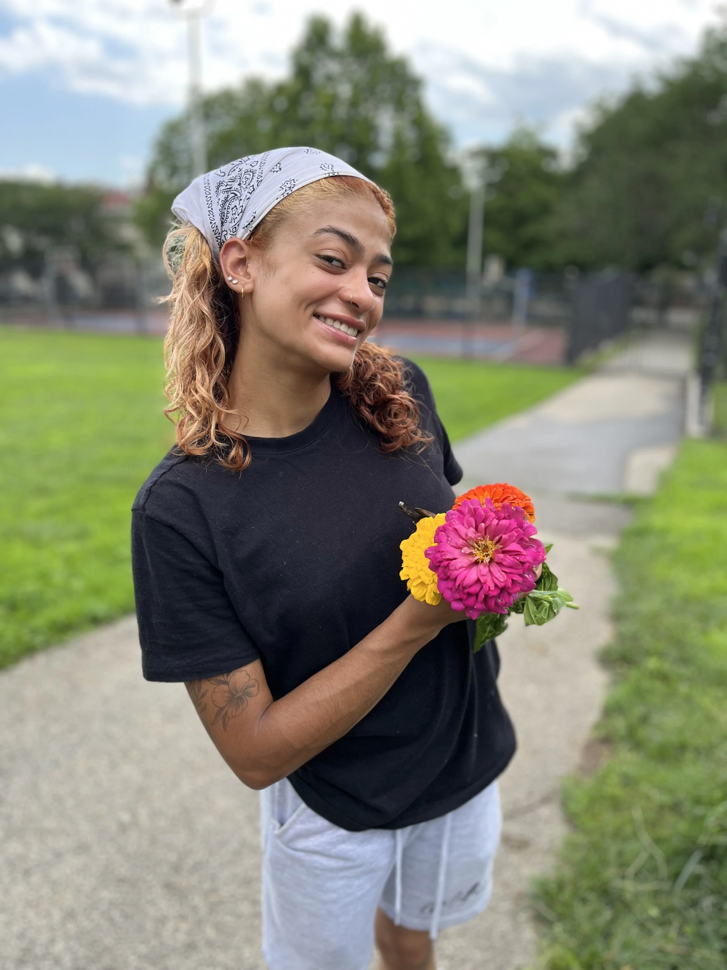 A young woman with curly hair, wearing a white bandana and a black t-shirt, holds a bunch of colorful flowers and stands on a park path.