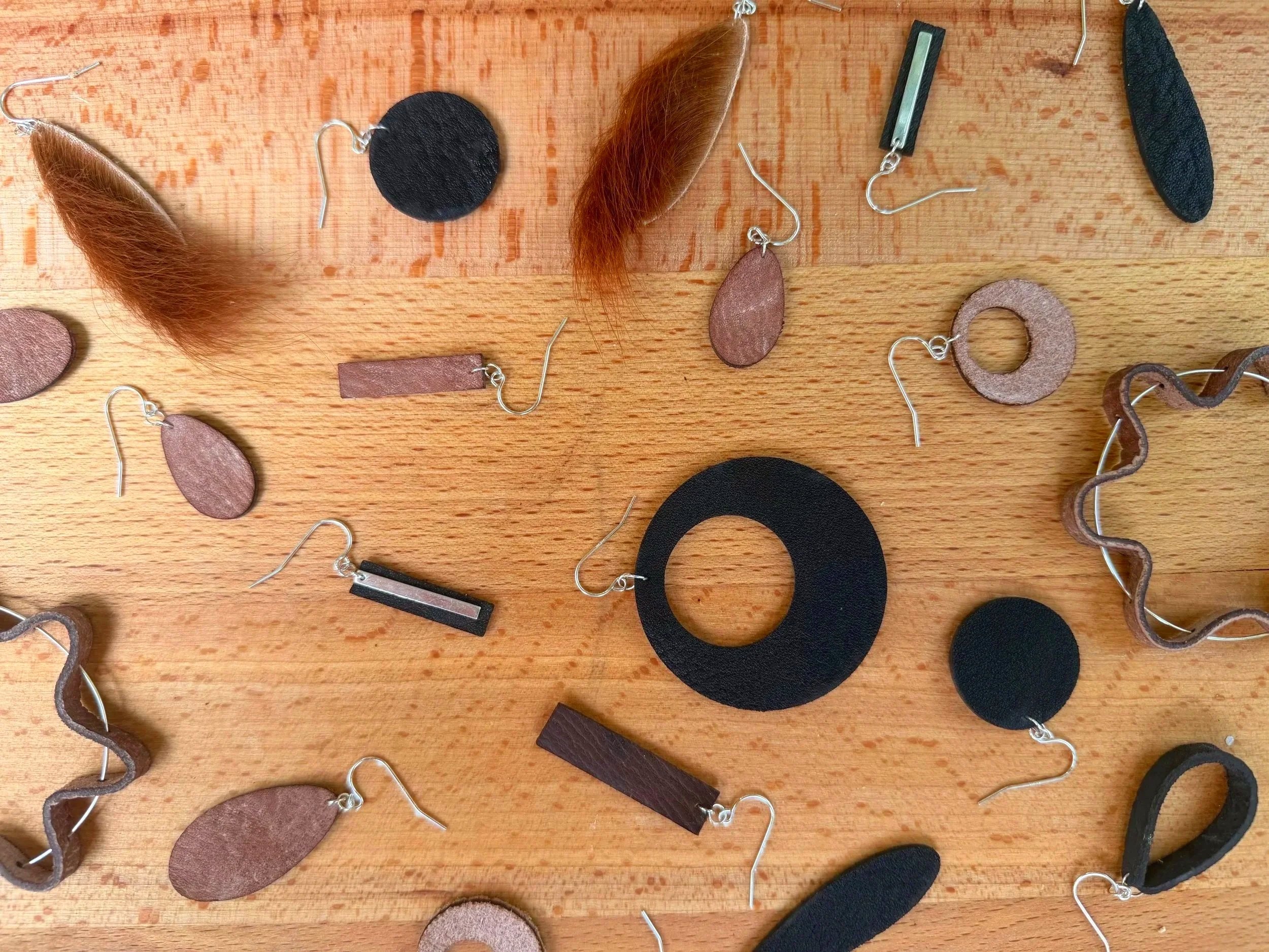 Assorted naturally tanned leather earrings in different shapes and finishes arranged on a wooden surface.