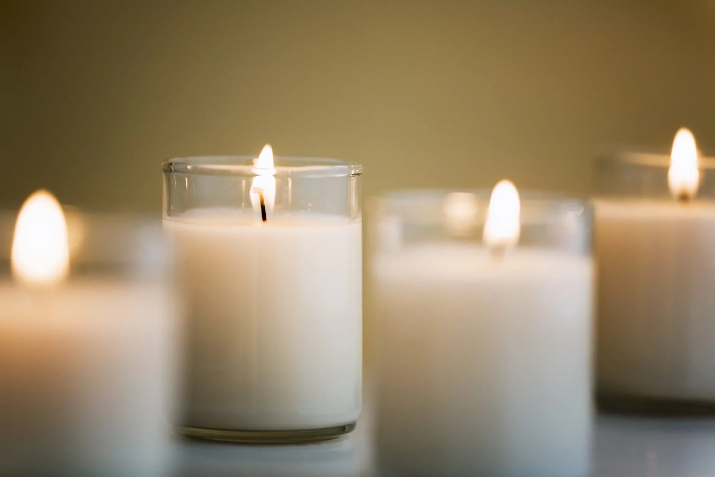 Close-up of lit white candles in glass holders on a smooth surface, with a plain, neutral-colored background.