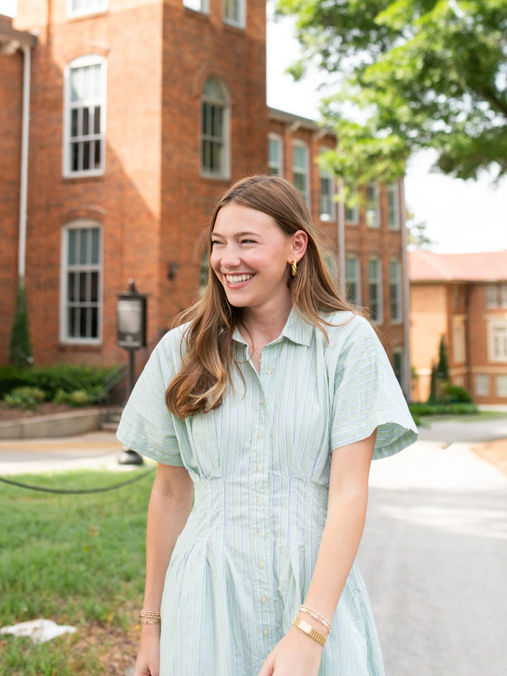 A young woman with long brown hair, wearing a light green striped dress, smiling outdoors near a brick building with trees.