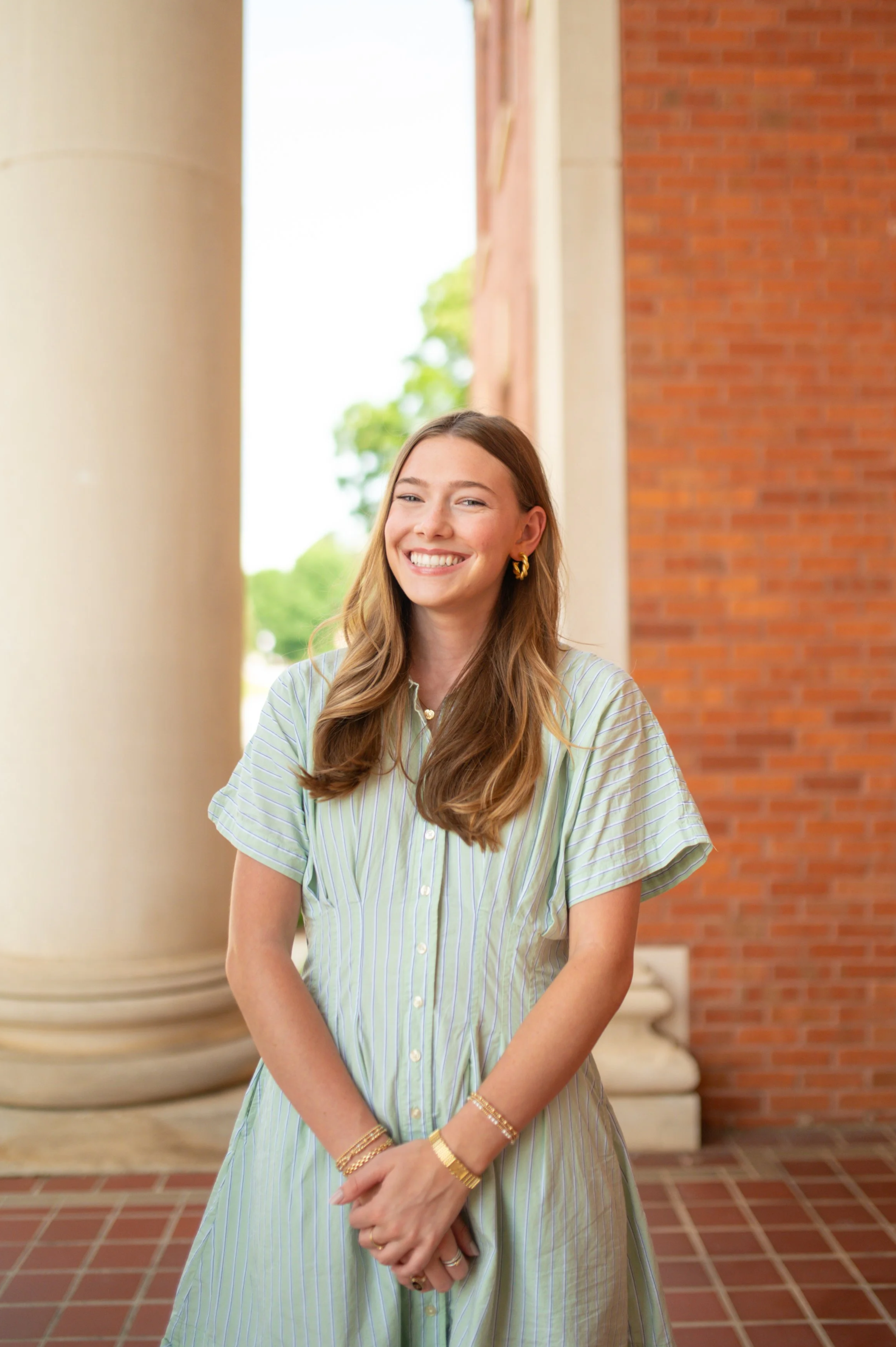 A young woman with long wavy hair smiling and wearing gold jewelry, standing outdoors in front of a brick building and large columns.
