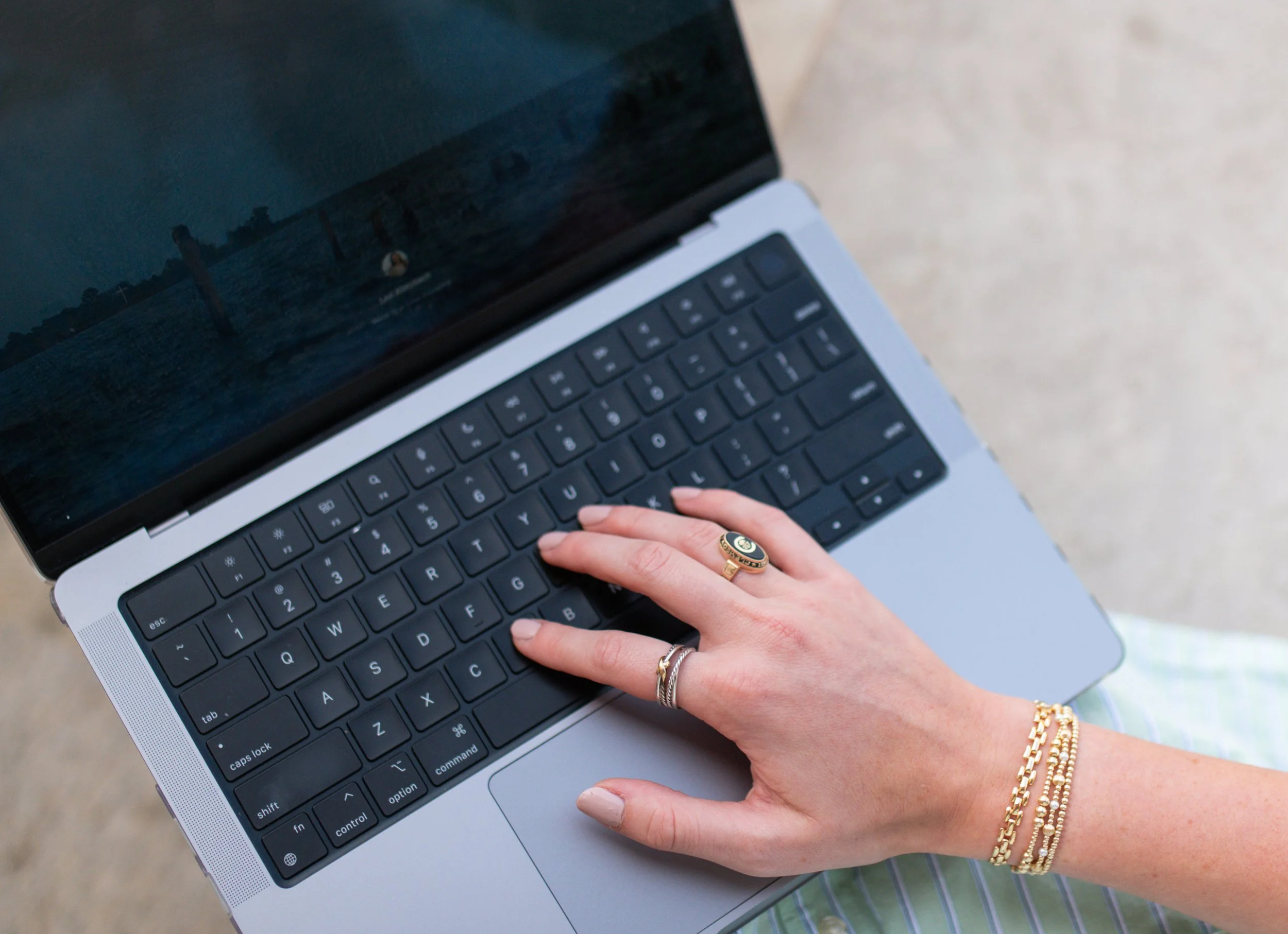 A person's hand with multiple gold rings and bracelets is resting on a silver MacBook keyboard, with a blurred background of an outdoor setting.
