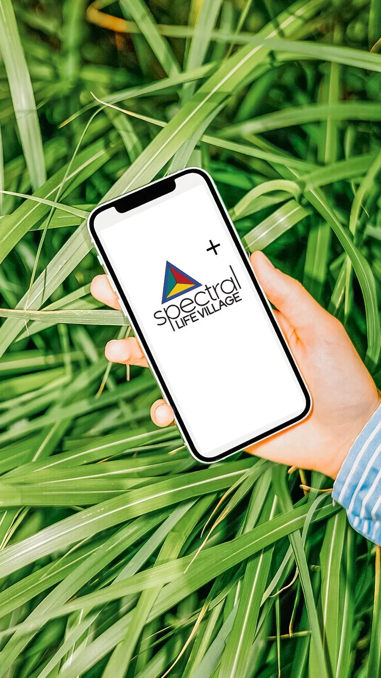 A person holding a smartphone with a logo that says 'spectral LIFE VILLAGE' and a colorful triangle, over green grass or leaves.