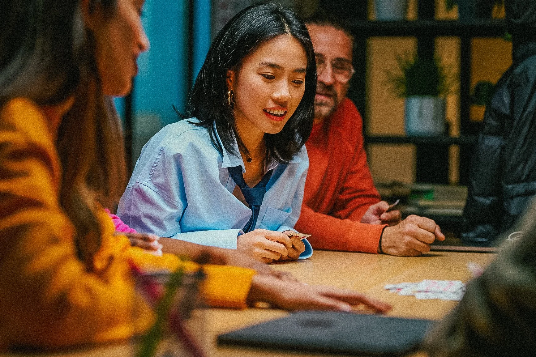 A group of people sitting at a table, engaged in a conversation or activity. The focus is on a woman with black hair, wearing a light blue shirt and a dark blue tie, smiling and holding some papers. The background shows a man with glasses and a red sweater and some office shelves with plants and books.