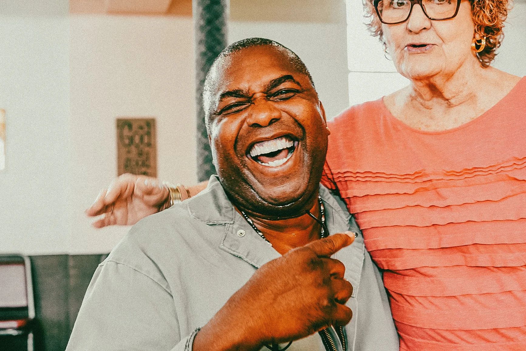A joyful man with dark skin, bald head, and big smile, being hugged by an older woman with light skin, curly hair, and glasses, in an indoor setting.