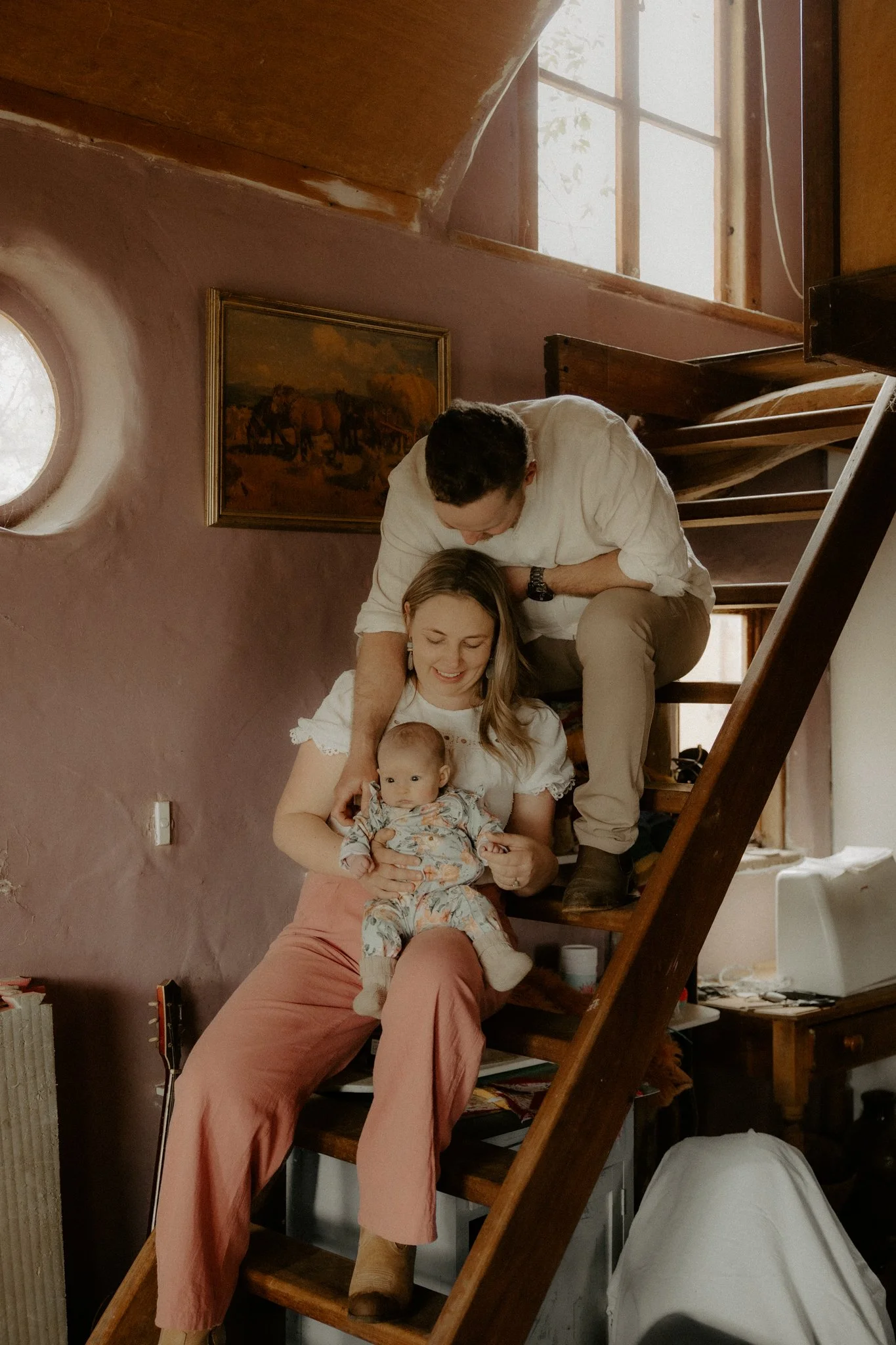 A family of three on a wooden staircase indoors. The mother, sitting on the stairs, holding a baby, with a man playfully leaning over her from above. The staircase is in a cozy home with pinkish walls, a painting on the wall, and a small round window