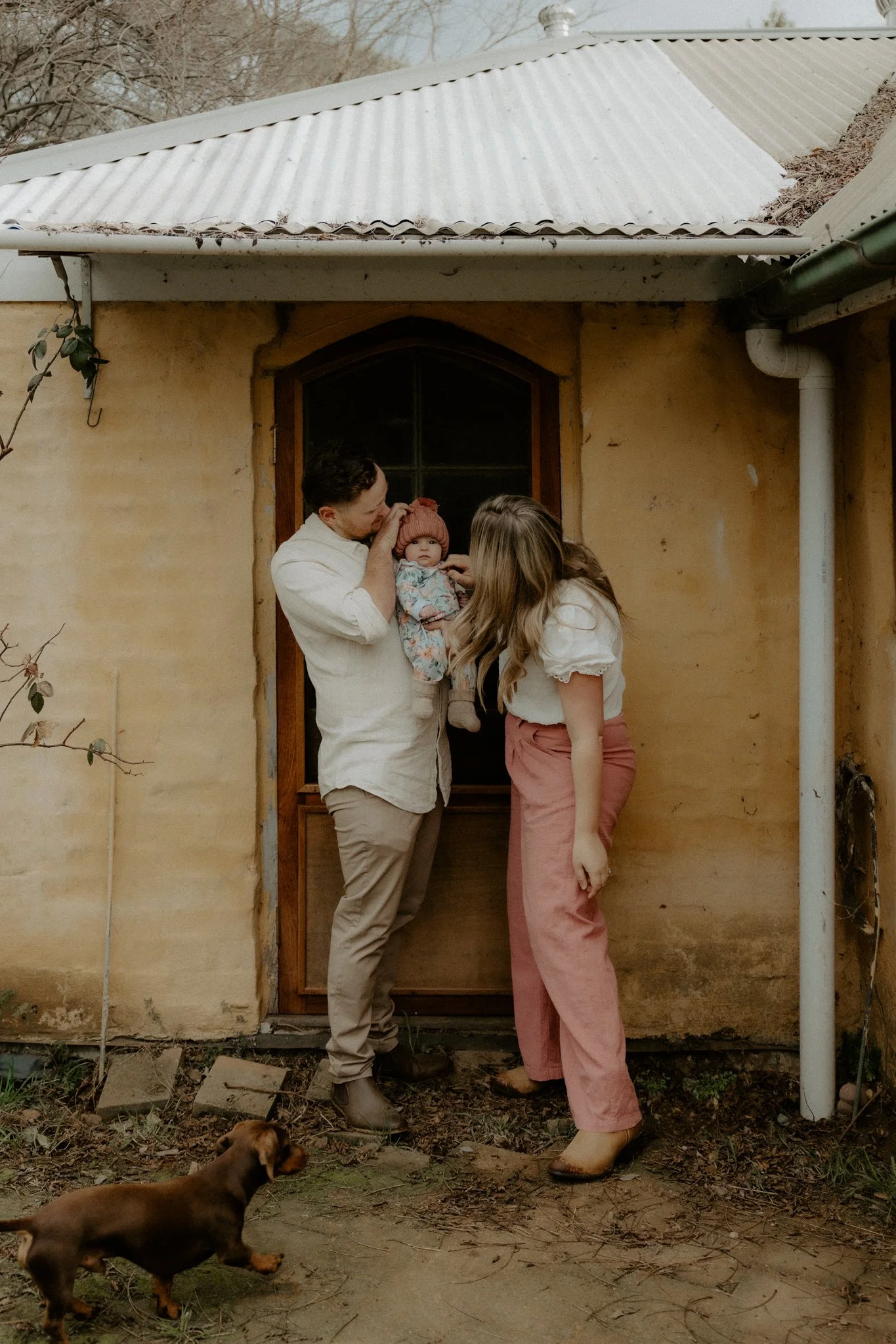 A family standing outside a rustic house, with a man holding a baby girl, a woman leaning in towards them, and a small brown dog in the yard.