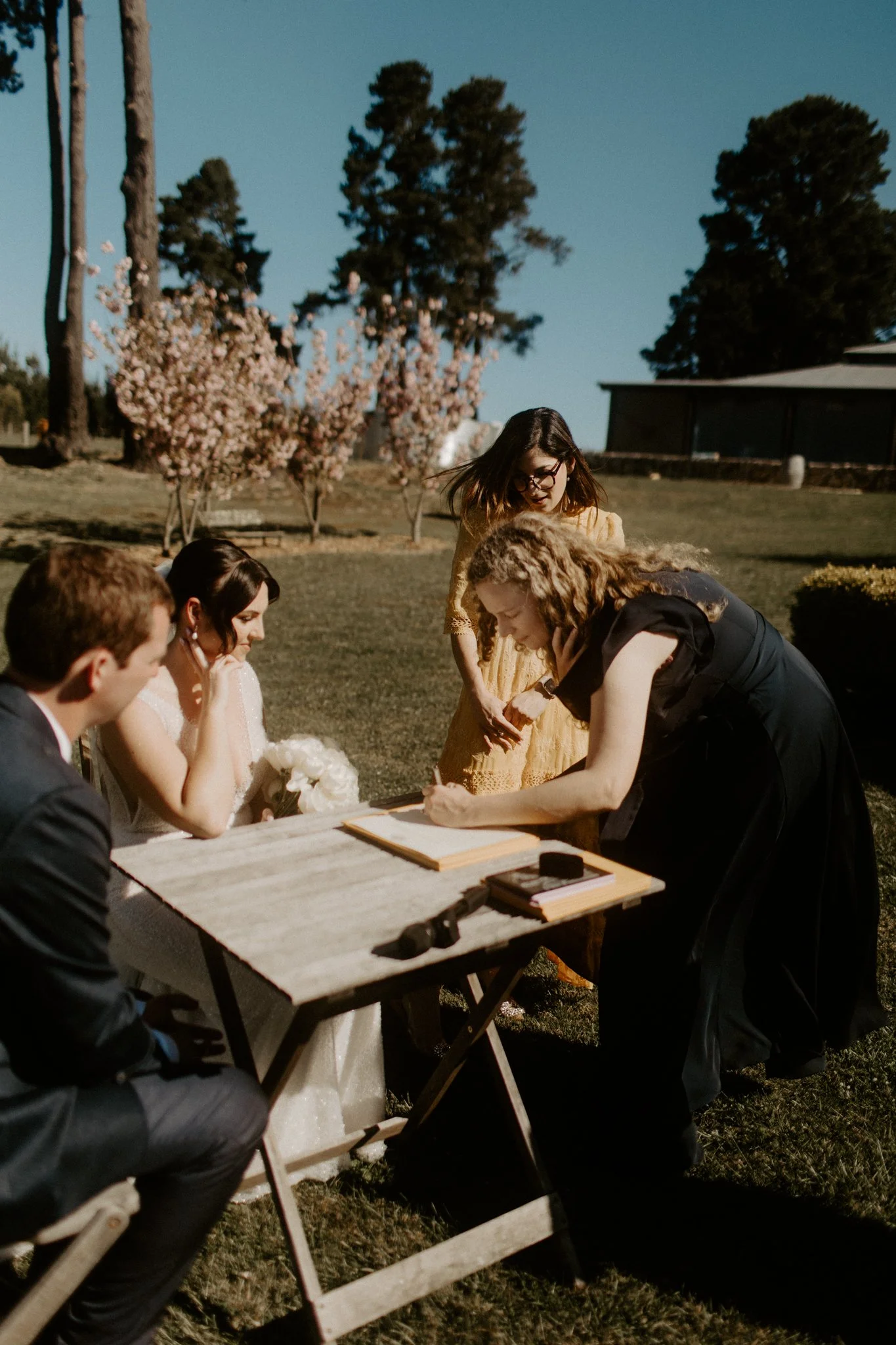 People gathered around a table outdoors, signing a document, during what appears to be a wedding ceremony on a sunny day with blooming trees in the background during a Blue Mountains wedding at Seclusions Blue Mountains.