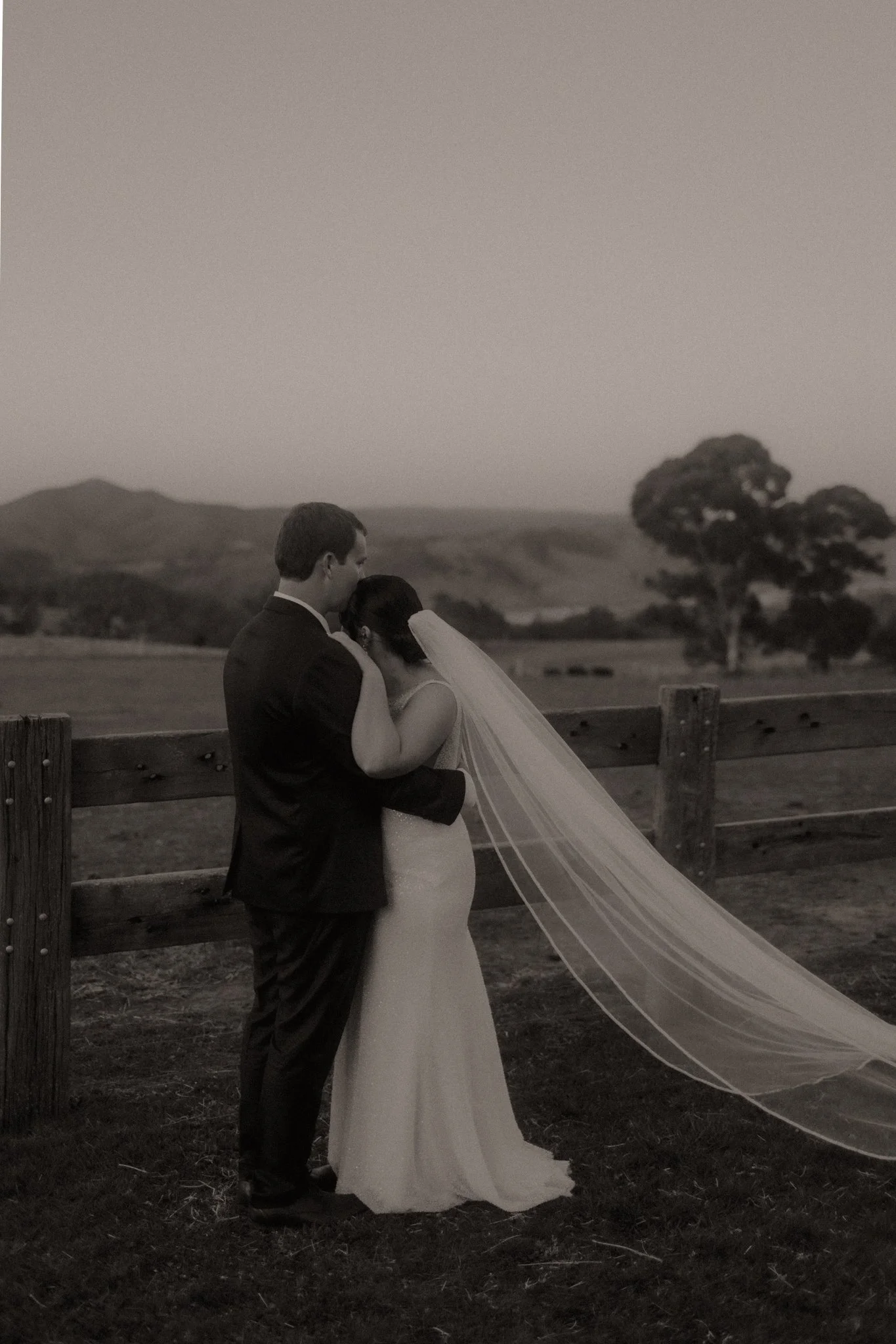 A black and white photo of a bride and groom embracing outdoors near a wooden fence, with rolling hills and a tree in the background.