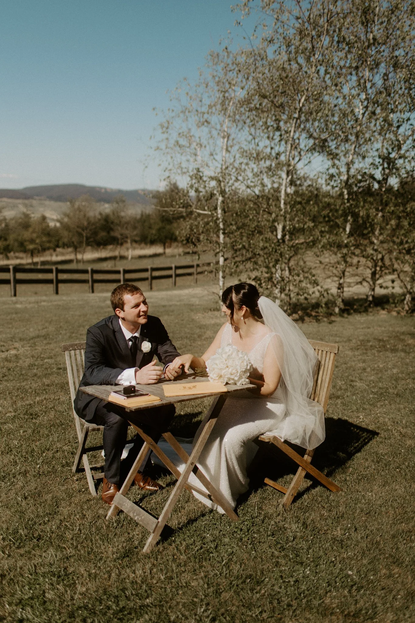 A bride and groom sitting at a rustic outdoor table on a grassy field with trees and hills in the background, during a Blue Mountains wedding at Seclusions Blue Mountains.