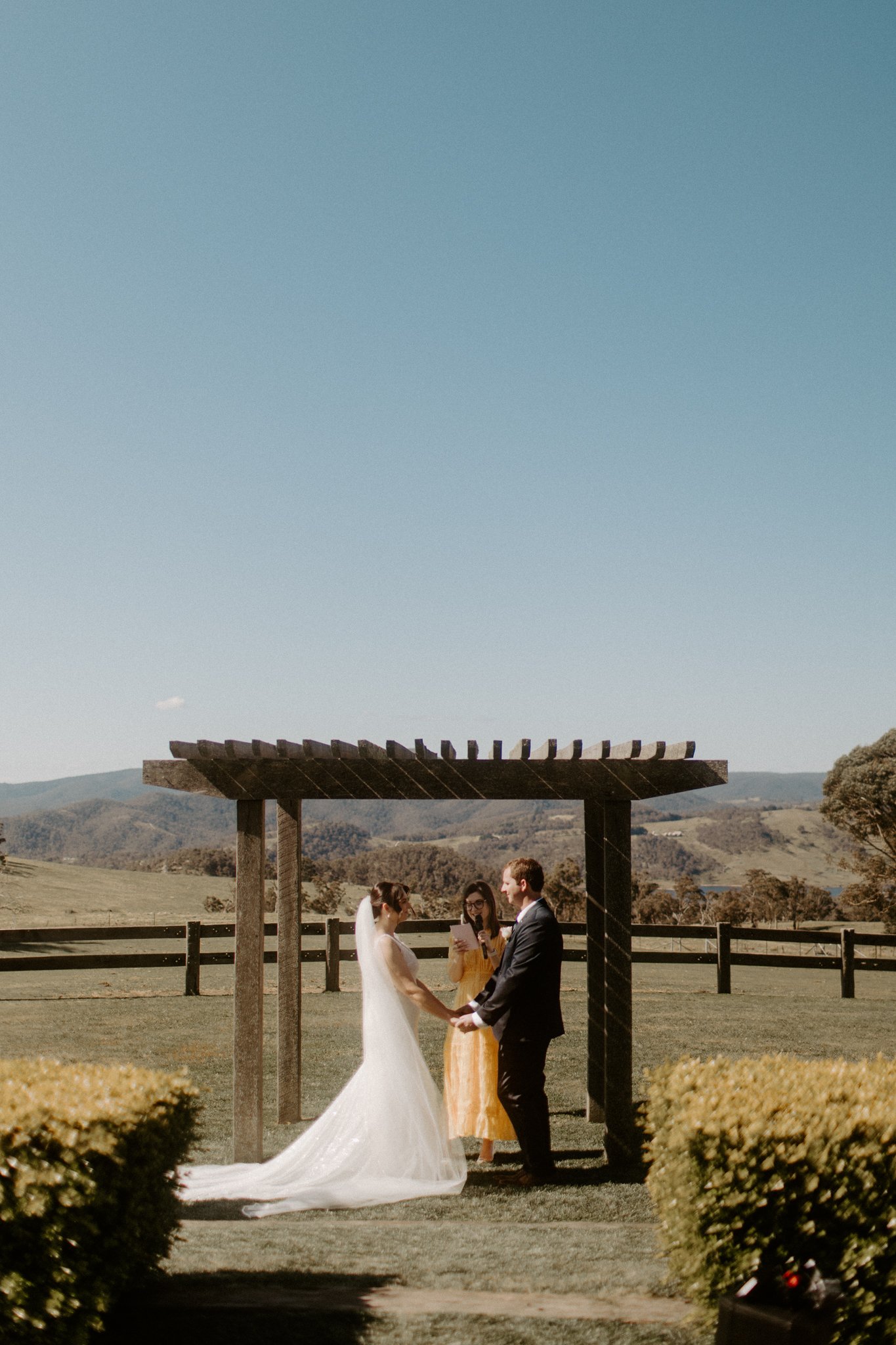 A wedding ceremony taking place outdoors with a bride and groom under a wooden arbor, in a scenic field with trees and mountains in the background at Seclusions Blue Mountains.