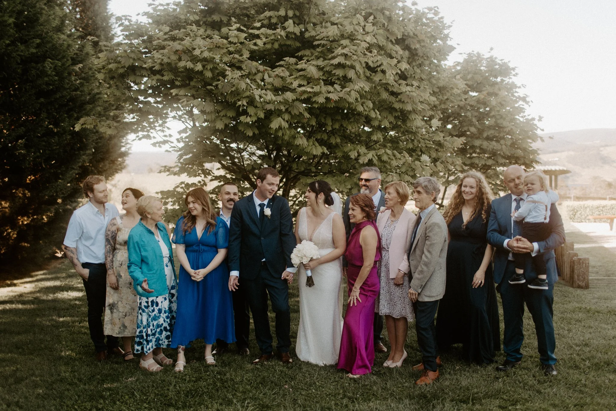 A group of people gathered outdoors for a wedding photo, including a bride and groom, family members, and friends, standing in front of a large leafy tree and green landscape.