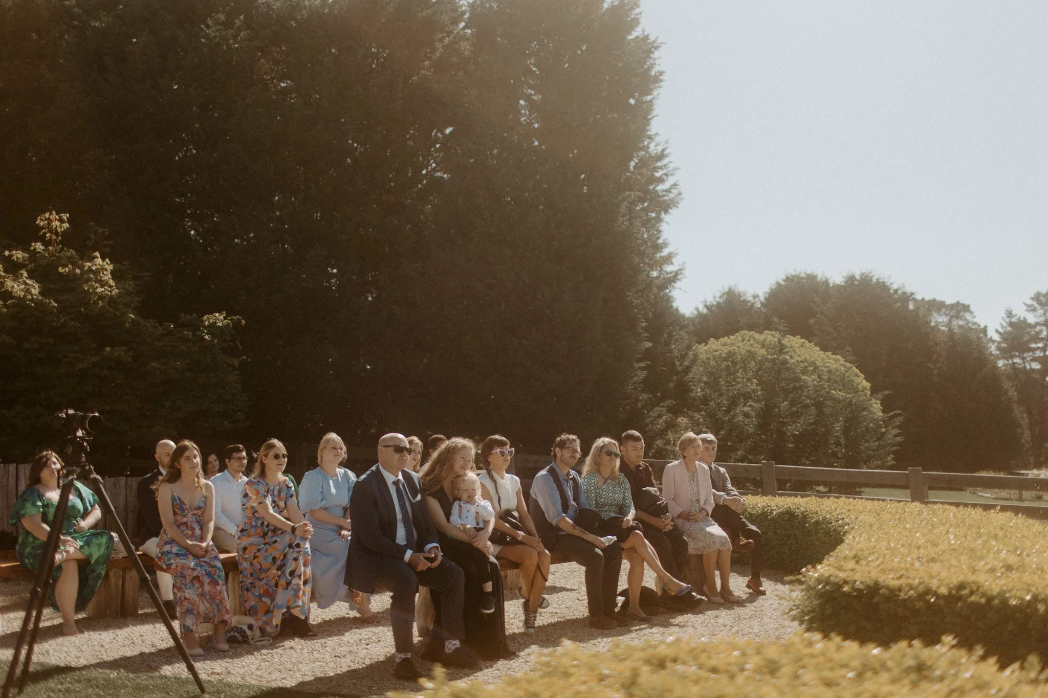 Group of people sitting outdoors on a sunny day, attending an event or ceremony, with trees and a wooden fence in the background, at Seclusions Blue Mountains.