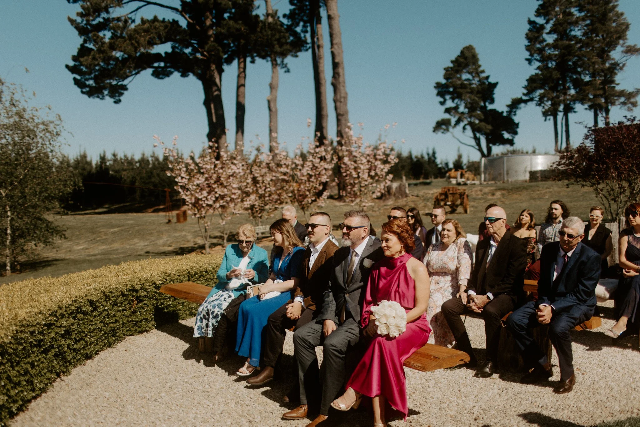 A group of people attending an outdoor wedding ceremony at Seclusions Blue Mountains, sitting on wooden benches, with trees and a blue sky in the background.