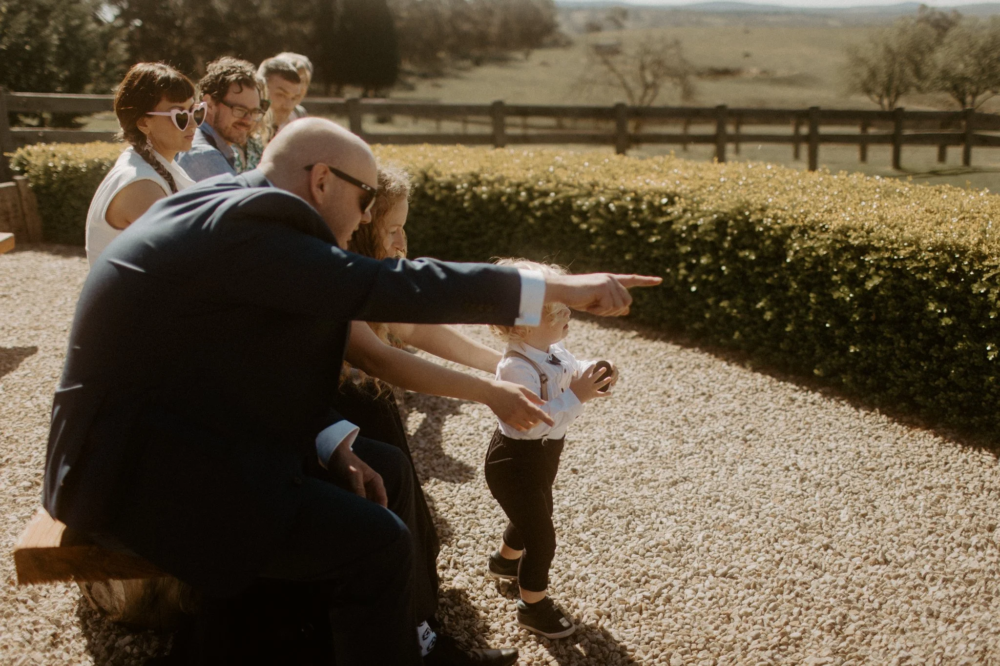 A group of people, including a man in a suit and a little , are seated outdoors on a wooden bench, appearing to watch and point at something in the distance on a bright sunny day. They are in a scenic, rural setting with a fence and fields in the bac