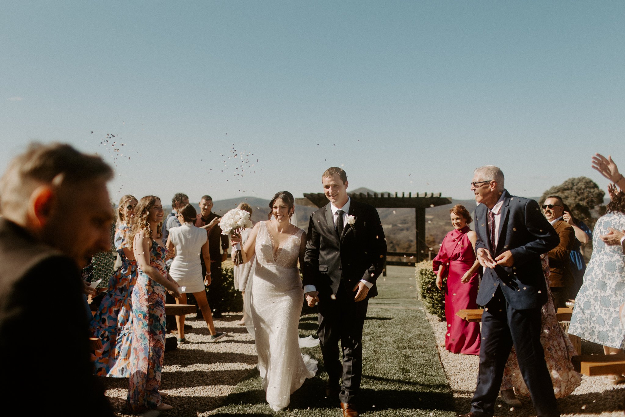A bride and groom walking down the outdoor aisle after their wedding ceremony, surrounded by smiling guests, some throwing confetti on a sunny day during a Blue Mountains wedding at Seclusions Blue Mountains.