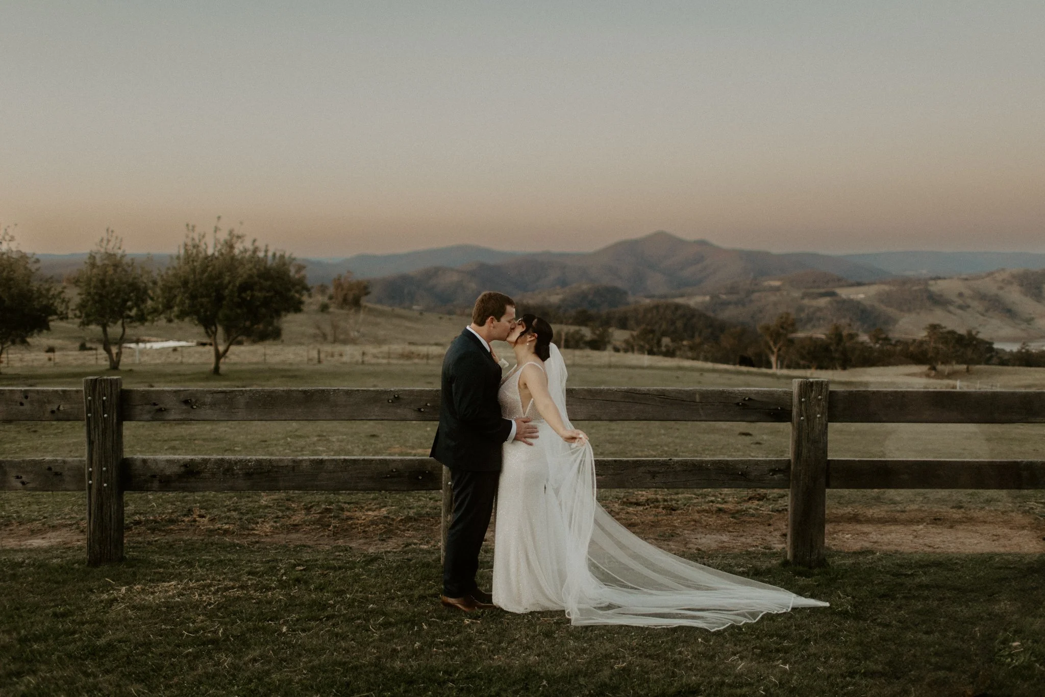 A bride and groom kissing outdoors in front of a wooden fence with a scenic landscape of rolling hills and trees at sunset.