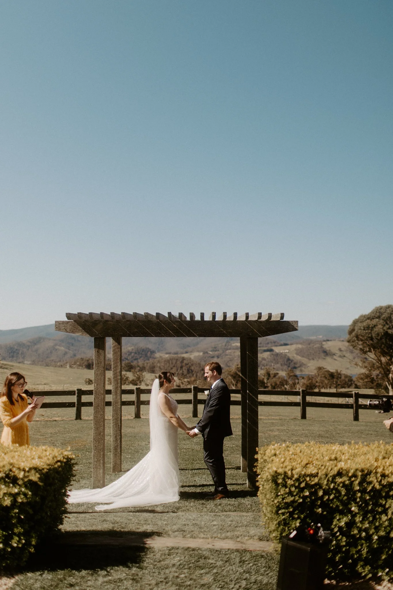 A wedding ceremony taking place outdoors with a bride and groom under a wooden arbor, in a scenic field with trees and mountains in the background at Seclusions Blue Mountains.