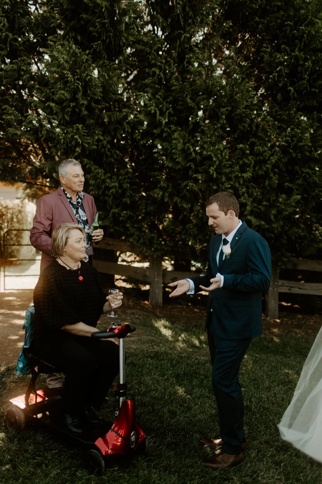 A young man in a suit giving a speech or saying something to a group of older adults outdoors, with people holding drinks, near a large tree, in a garden or backyard setting.