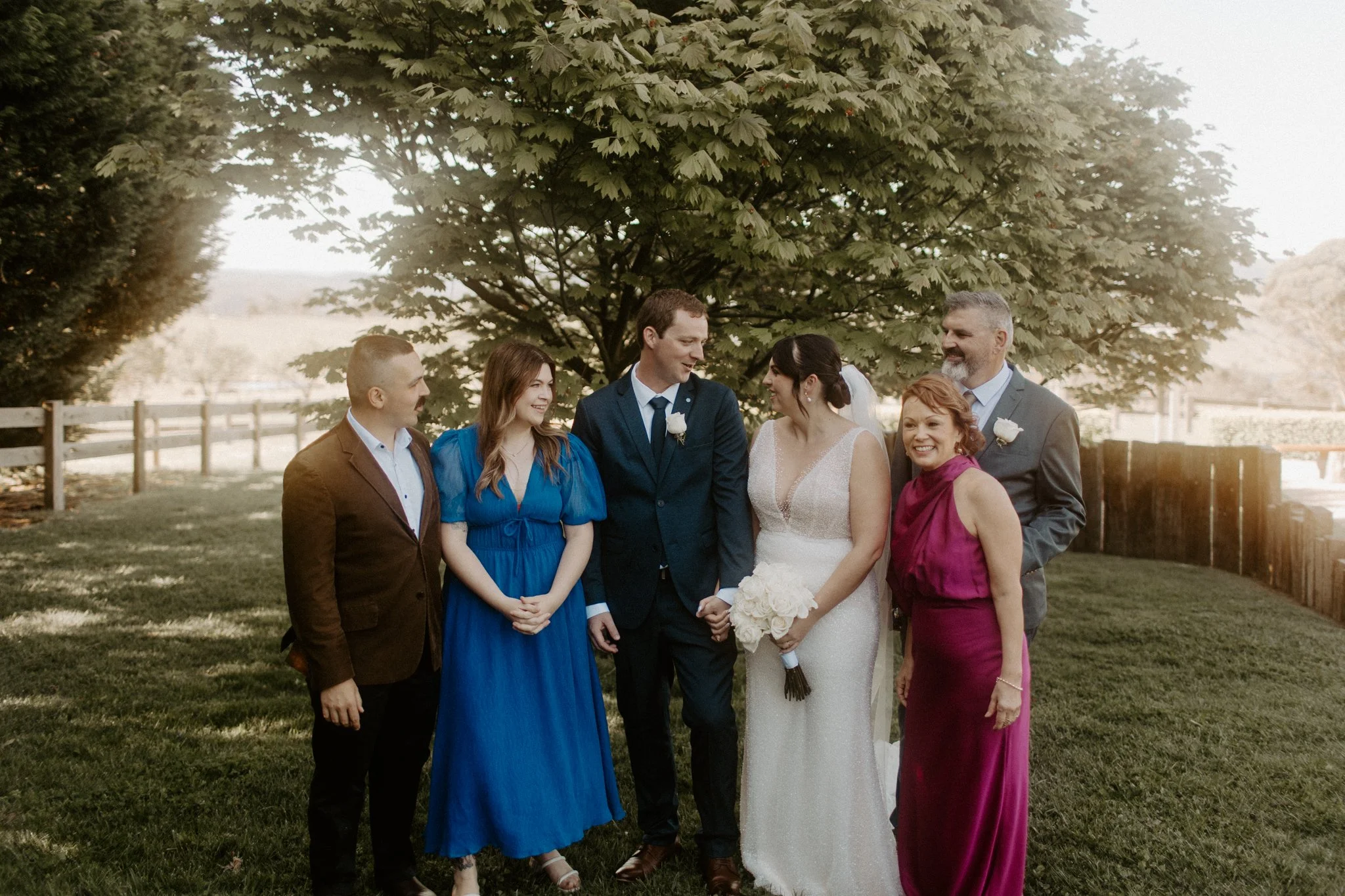 A wedding group photo outdoors under a tree, including a bride in a white gown holding a bouquet, the groom in a navy suit, and four other people in colorful dresses and suits.
