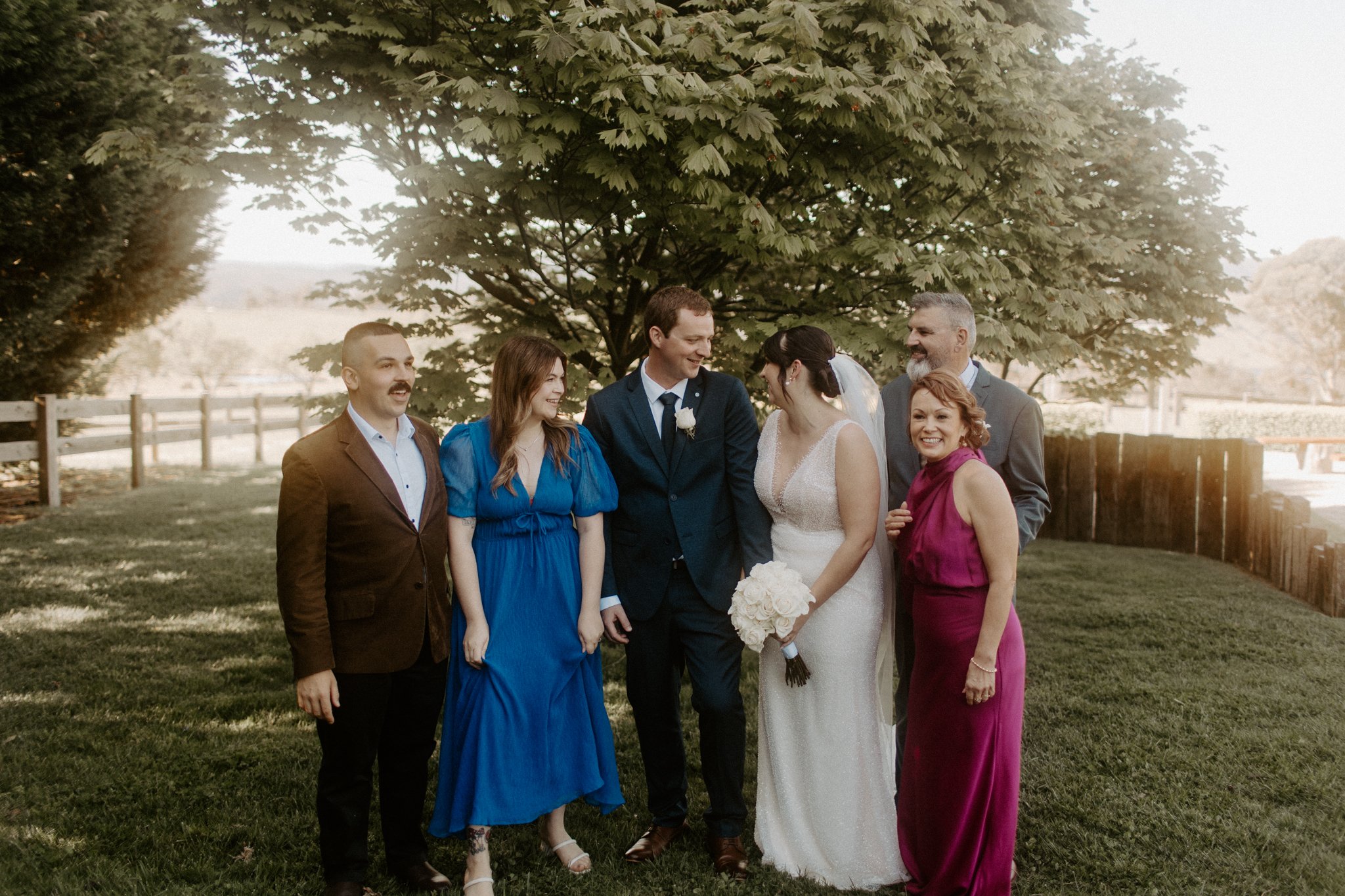 A group of six people, including a bride in a white wedding dress holding a bouquet of white flowers and a groom in a dark suit, standing outdoors under a large tree on a sunny day, smiling and looking at each other. The others are dressed in semi-fo