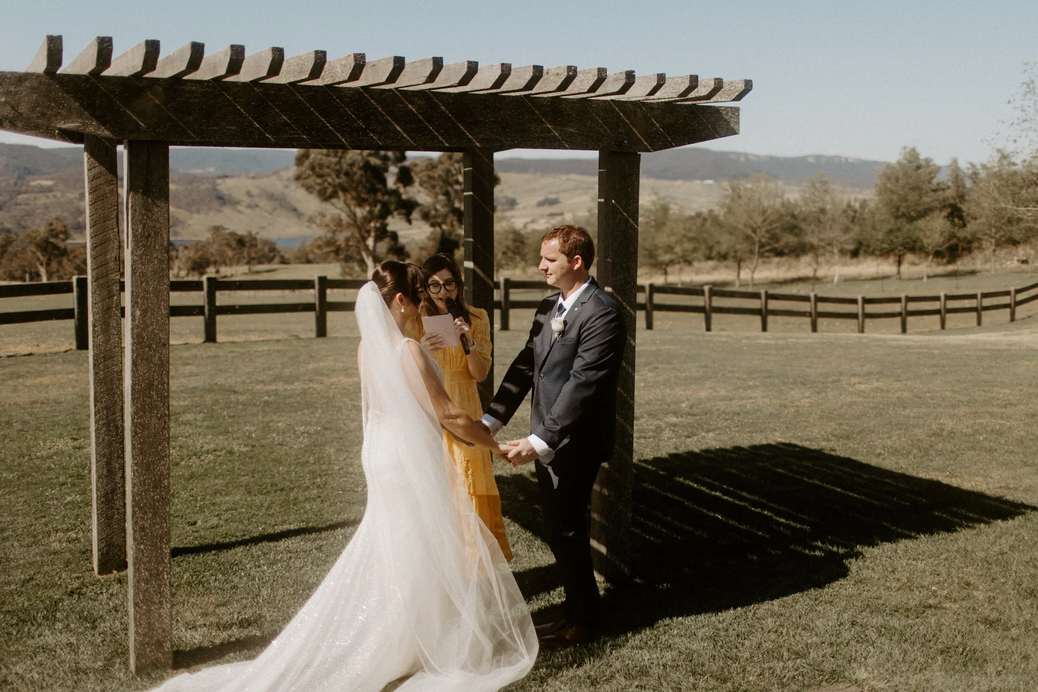 A wedding ceremony taking place outdoors with a bride and groom under a wooden arbor, in a scenic field with trees and mountains in the background at Seclusions Blue Mountains.