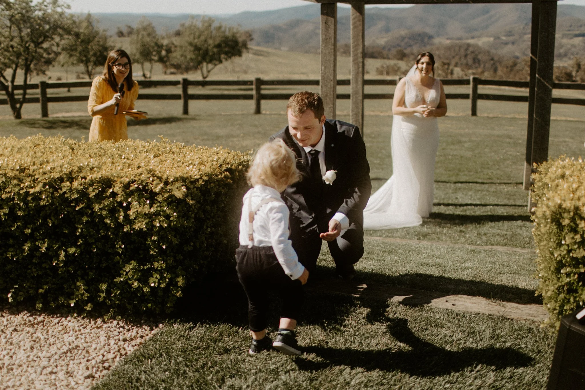 A groom kneeling and talking to a young boy during an outdoor wedding ceremony, with a bride in a white wedding dress smiling in the background during a Blue Mountains wedding at Seclusions Blue Mountains.