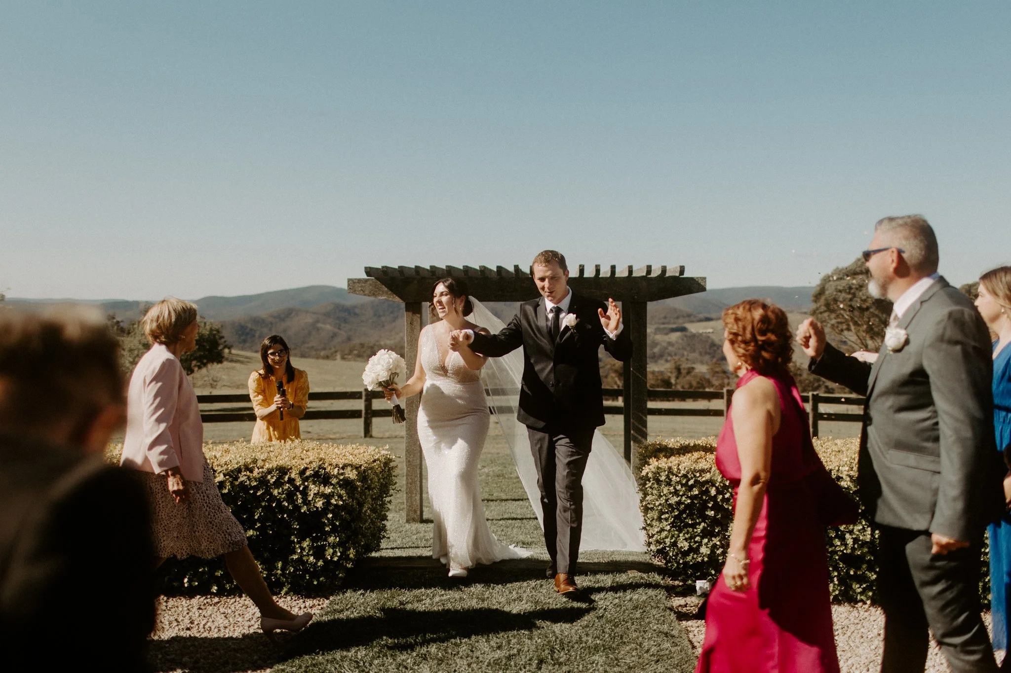 A bride and groom walking down the aisle outdoors with guests celebrating around them, scenic mountains in the background during a Blue Mountains wedding at Seclusions Blue Mountains.