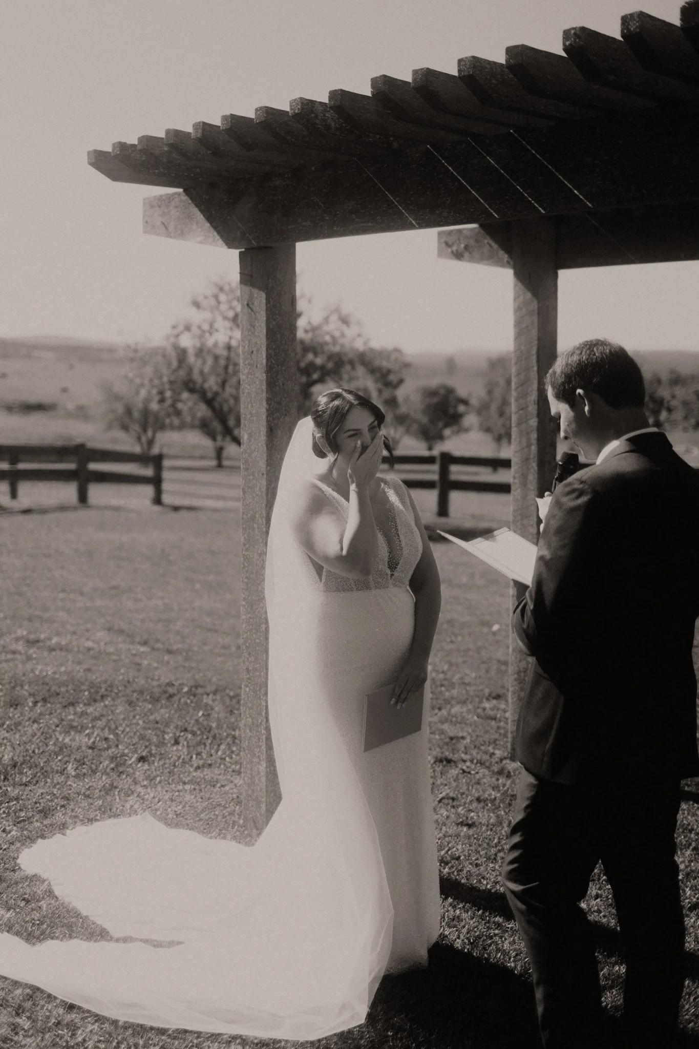 A wedding ceremony taking place outdoors with a bride and groom under a wooden arbor, in a scenic field with trees and mountains in the background at Seclusions Blue Mountains.