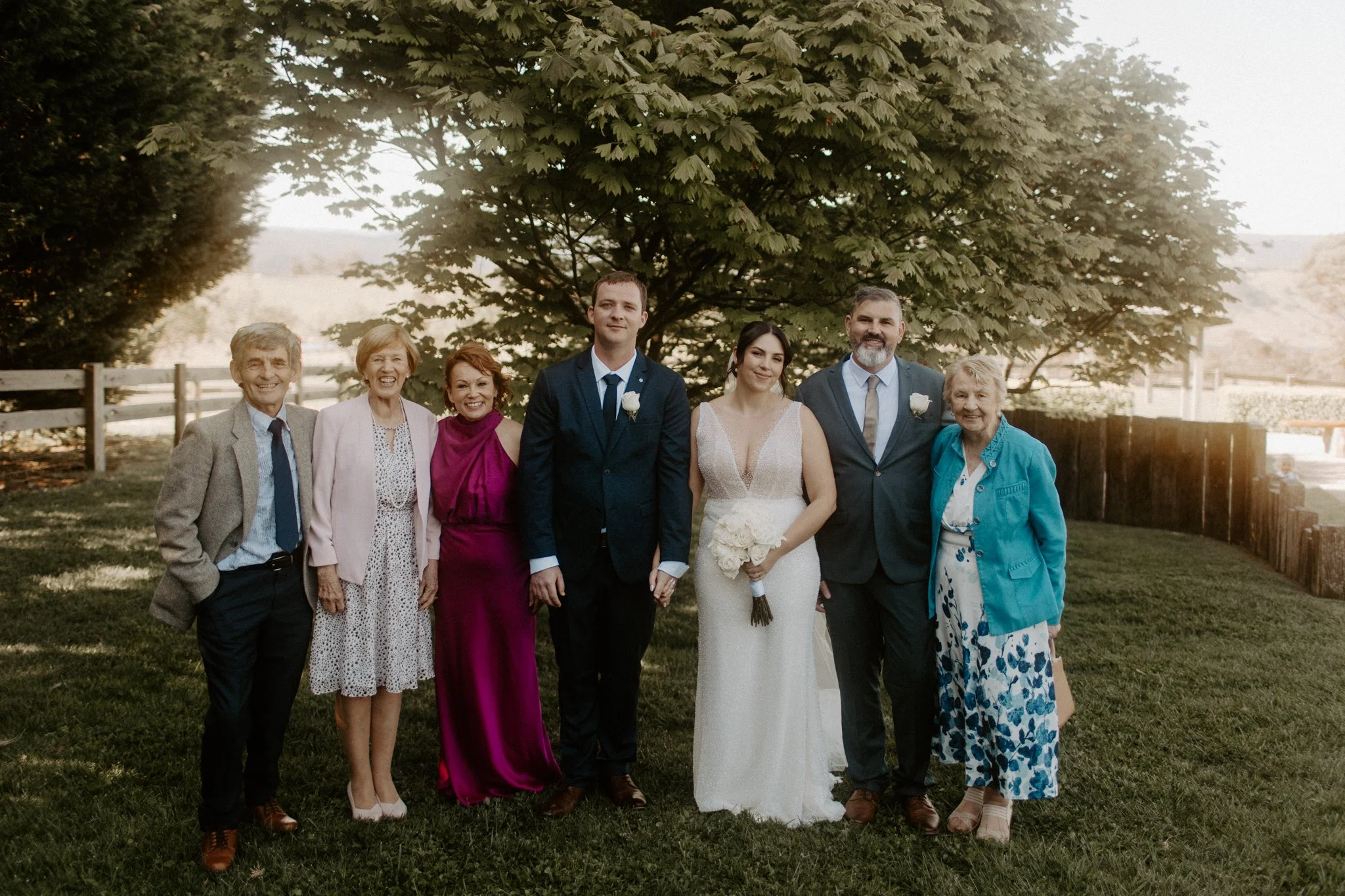 Group of people standing outdoors in front of a tree, at a wedding or celebration, dressed in formal attire.
