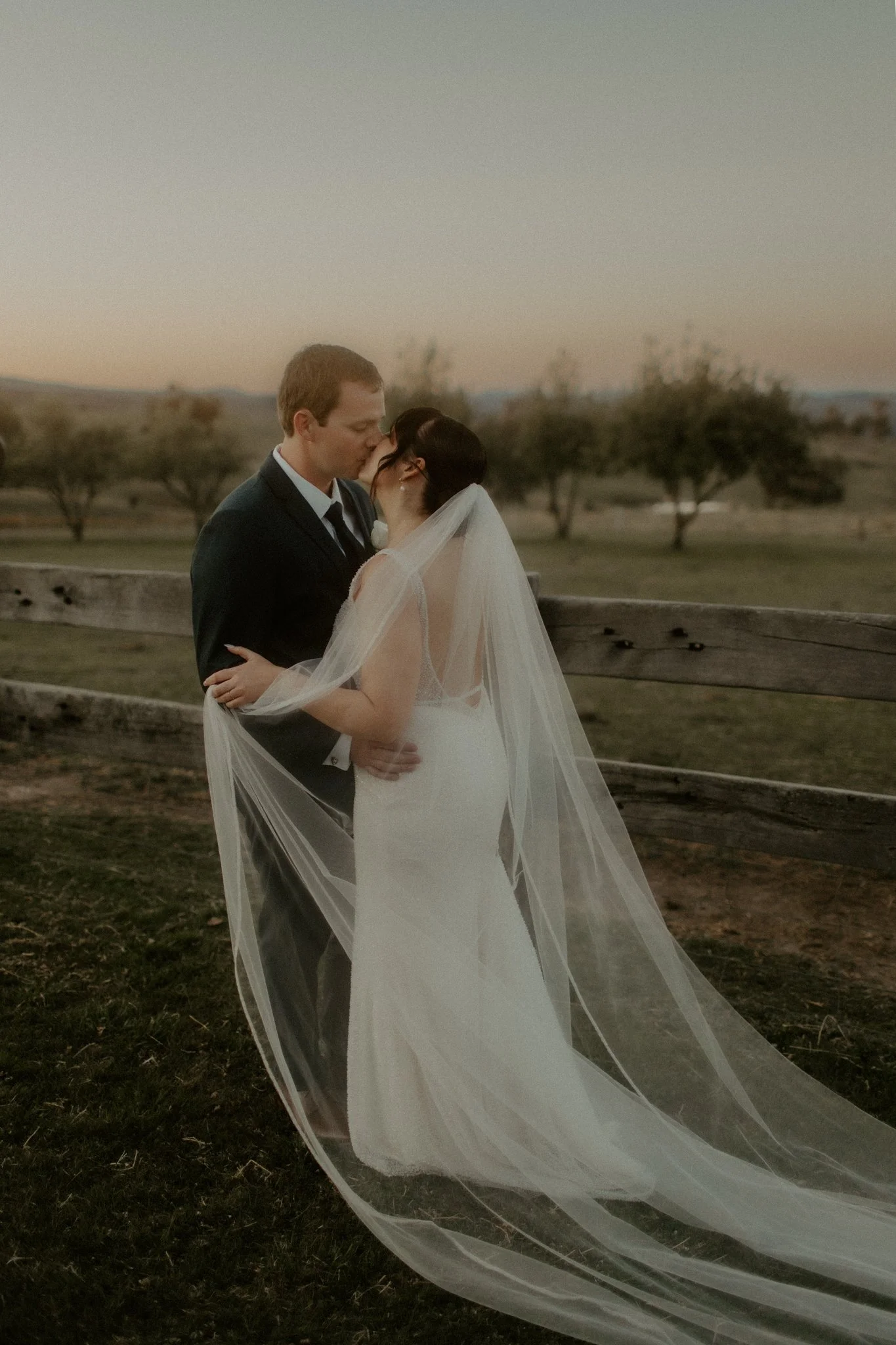 A bride and groom sharing a kiss outdoors near a wooden fence during sunset, with trees and a landscape in the background.