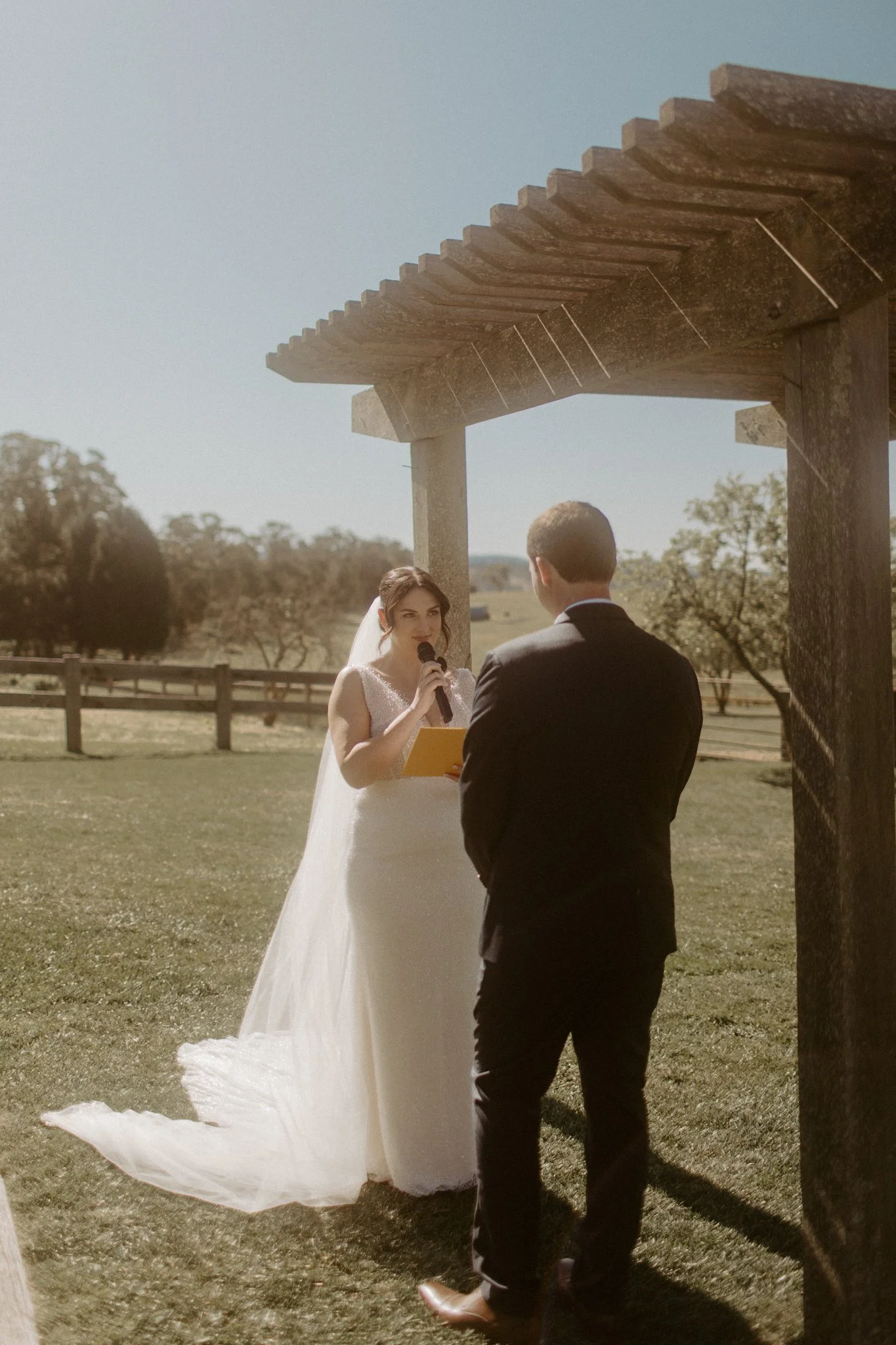 A wedding ceremony taking place outdoors with a bride and groom under a wooden arbor, in a scenic field with trees and mountains in the background at Seclusions Blue Mountains.