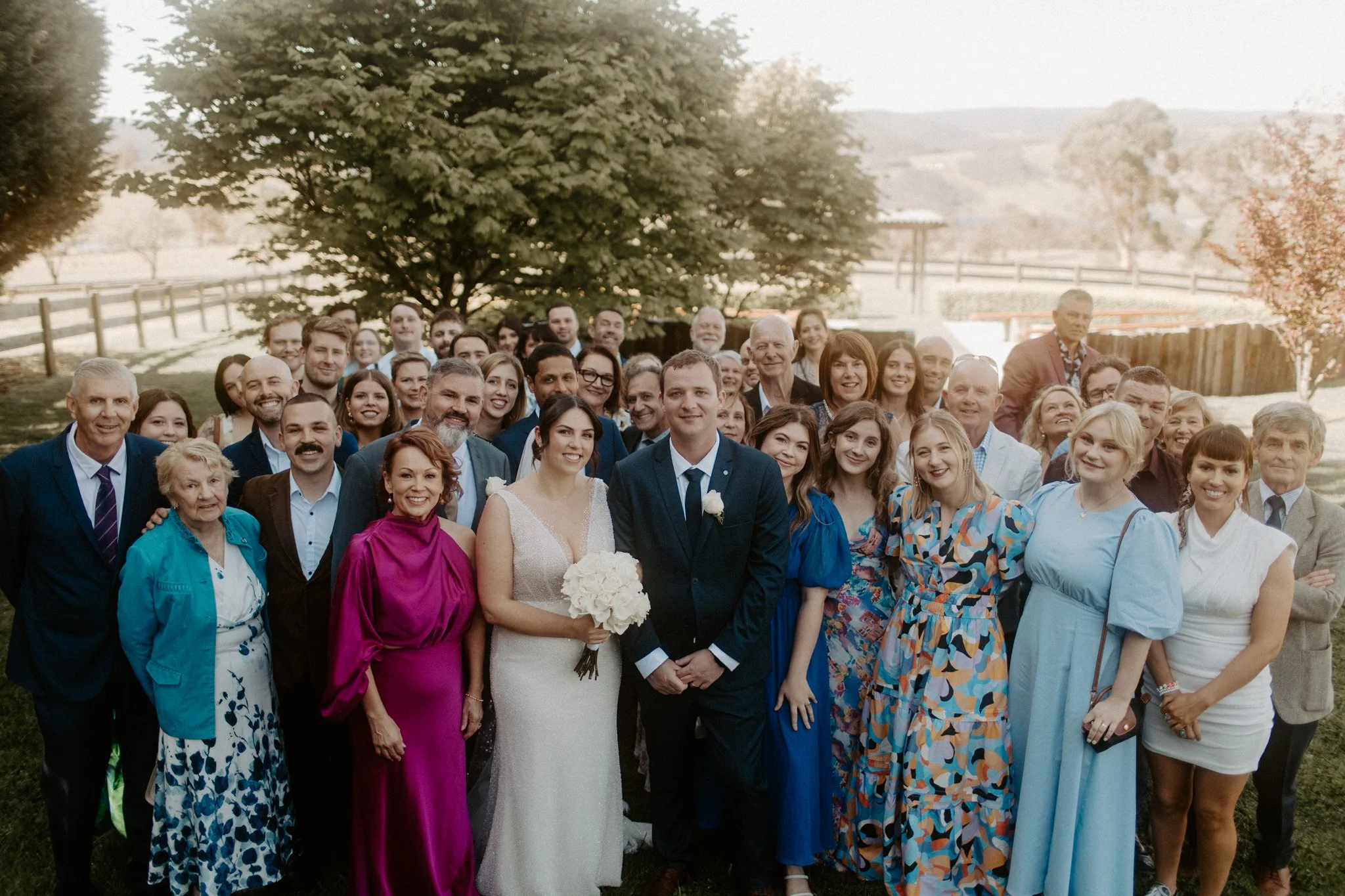 A large group of people celebrating outdoors at a wedding, including the bride in a white gown holding a bouquet, the groom in a dark suit, and guests in colorful dresses and suits, with trees and a wooden fence in the background.