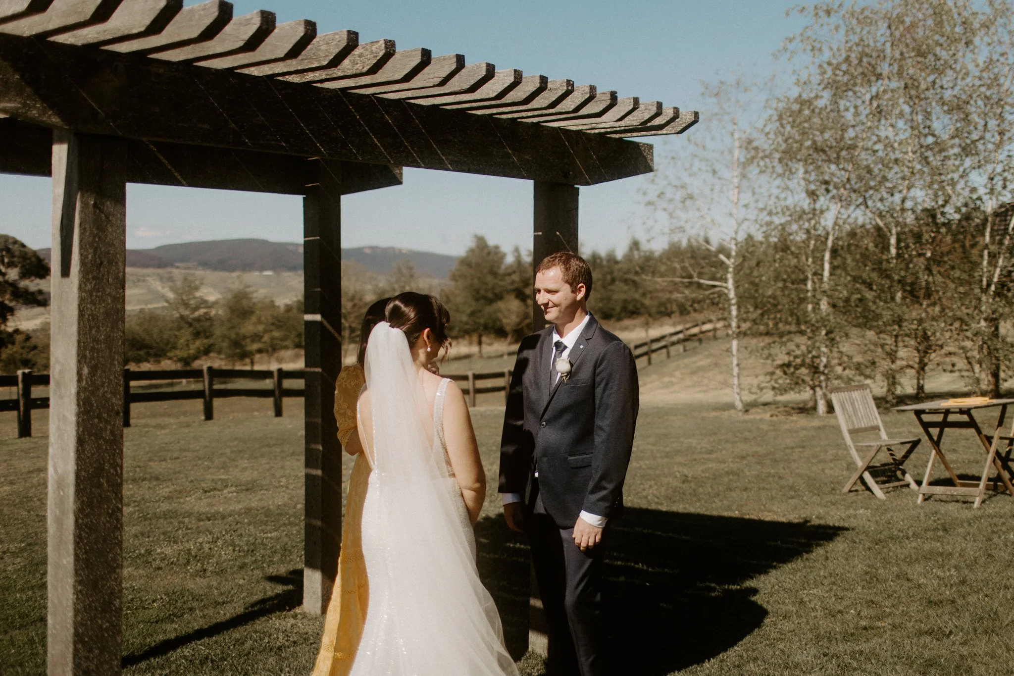 A wedding ceremony taking place outdoors with a bride and groom under a wooden arbor, in a scenic field with trees and mountains in the background at Seclusions Blue Mountains.