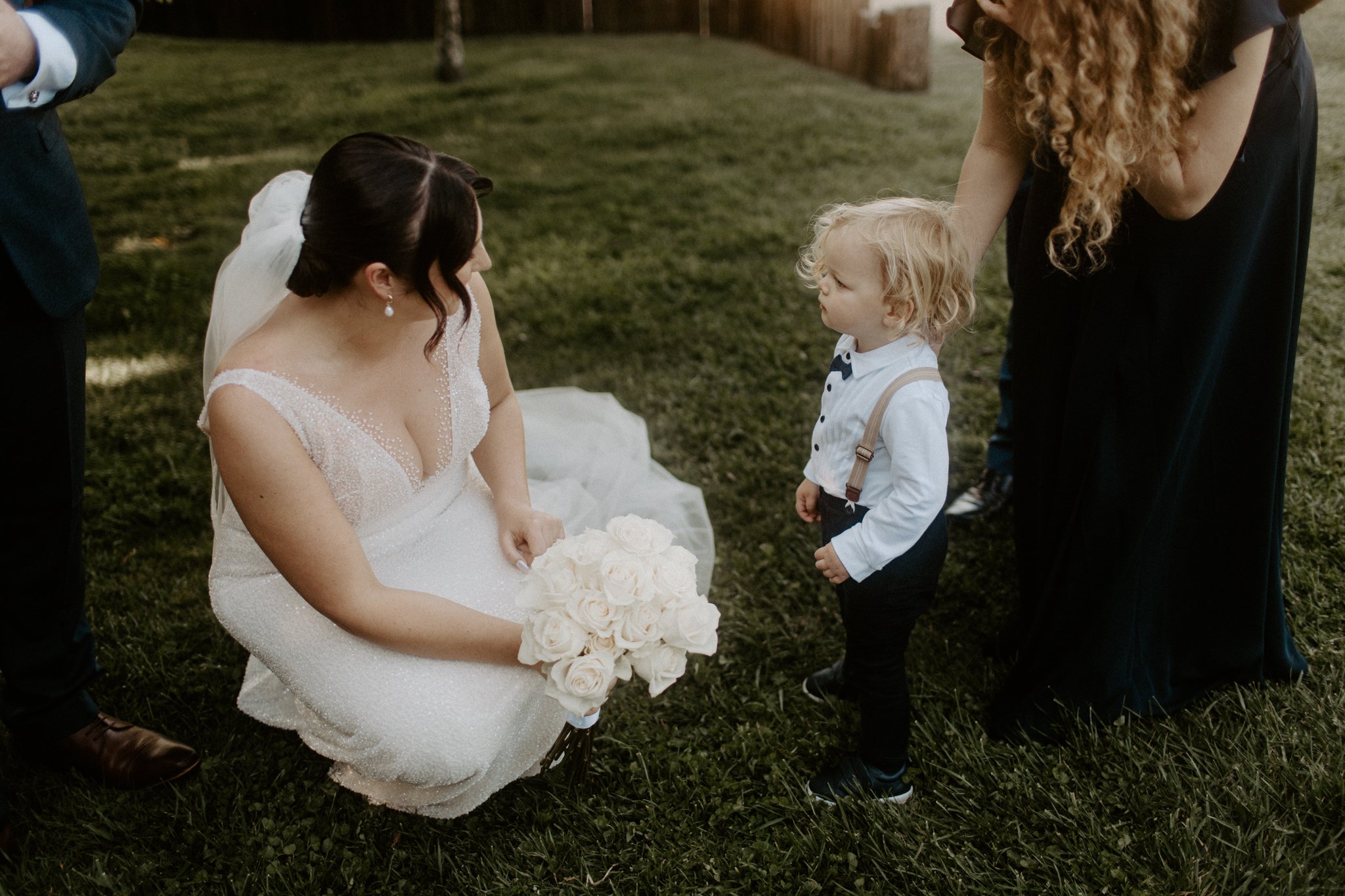 A bride in a white wedding dress kneeling on grass and holding a bouquet of white roses, looking at a young boy with blond hair, dressed in a white shirt with suspenders, who is standing and looking back at her. An adult woman with long curly red hai