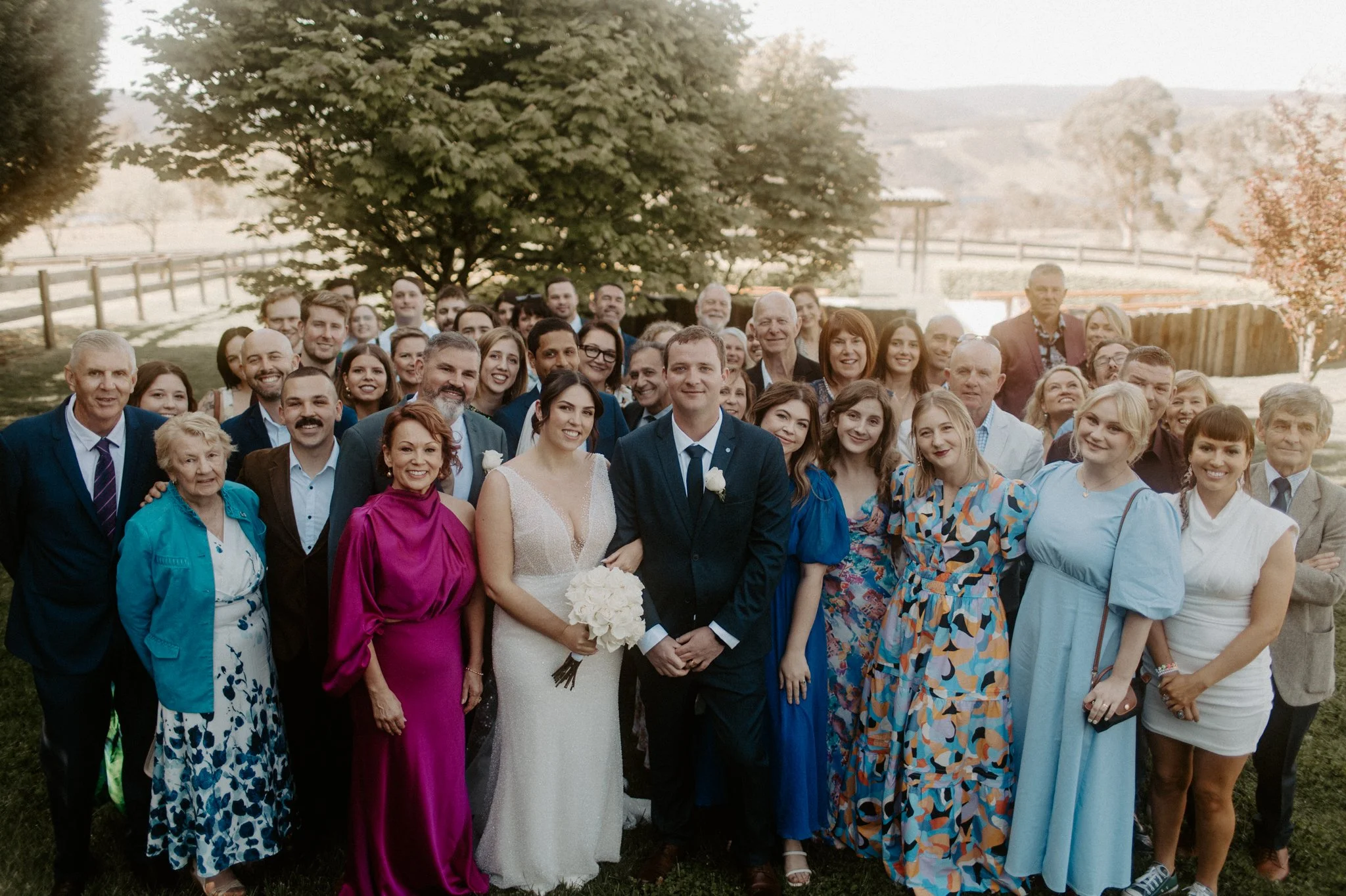 A large group of people, including a bride and groom, standing outdoors under a large tree, celebrating a wedding.