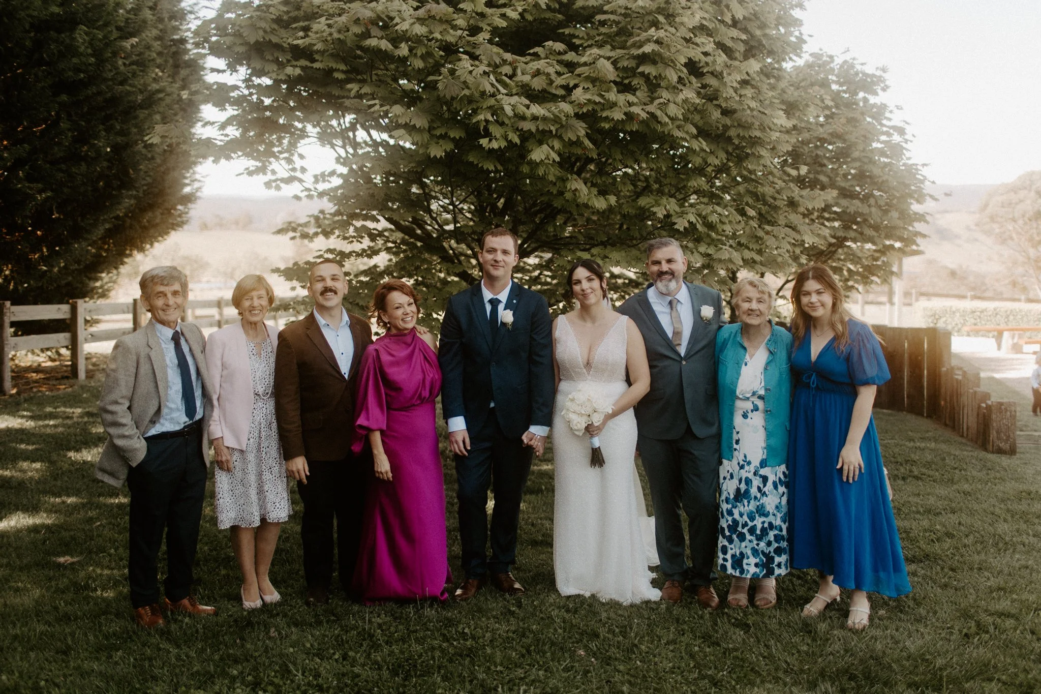 Group of nine people standing outdoors under a large tree during a wedding, with a bride and groom in the center, dressed in wedding attire, surrounded by family and friends.