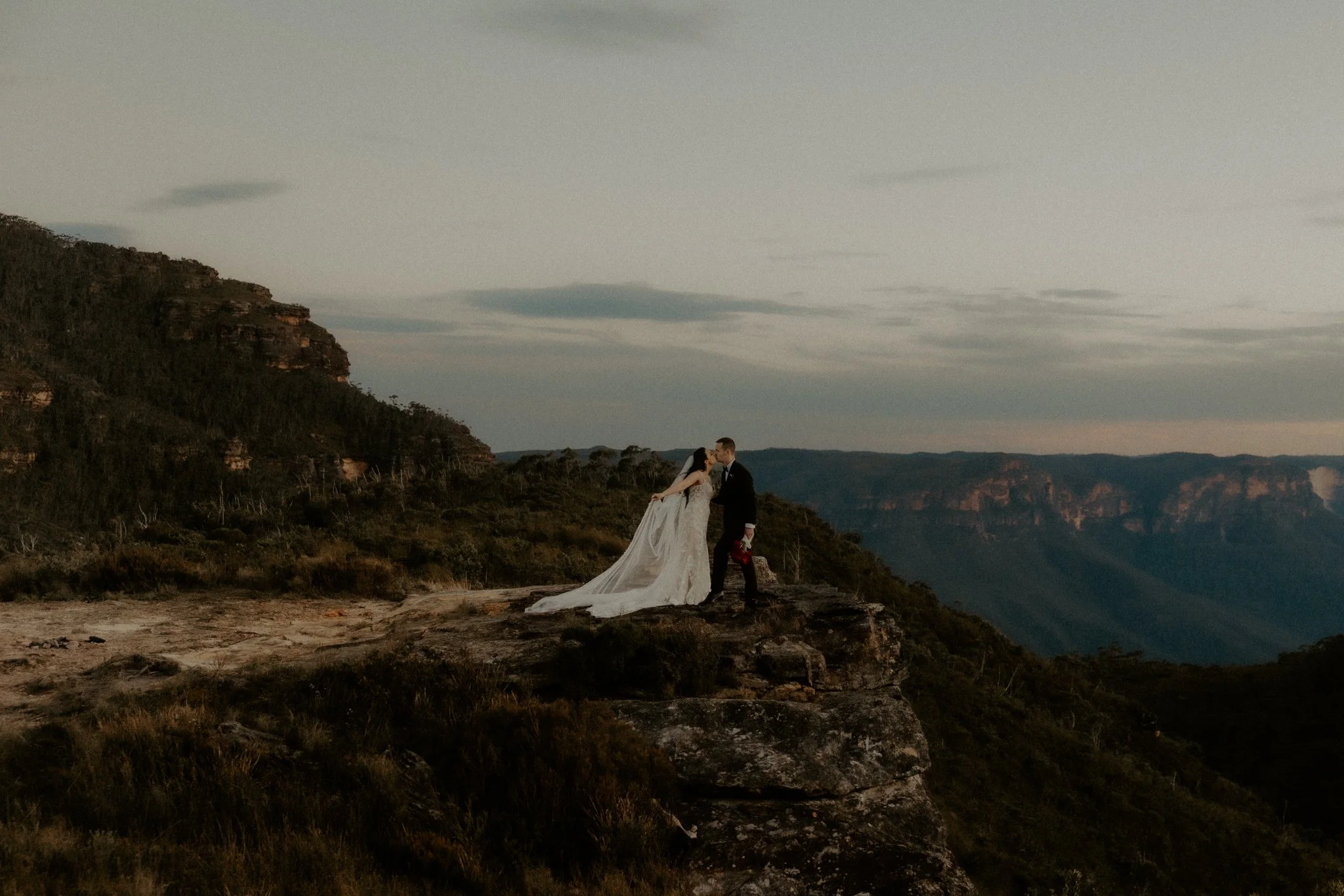 A bride and groom kissing on a rocky cliff with a mountainous landscape in the background at sunset.