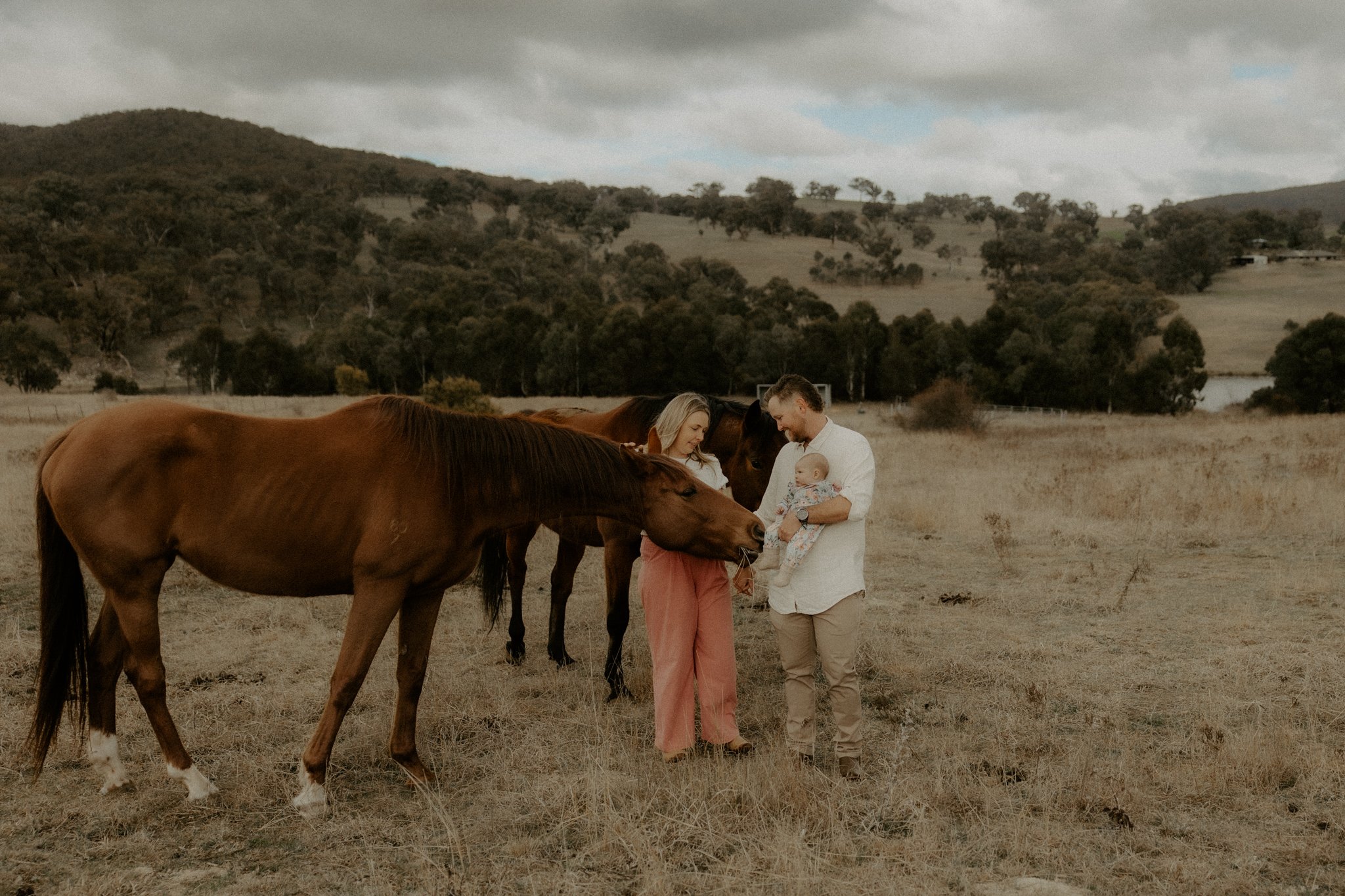 Family with two horses in an open field with hills and trees in the background, overcast sky.