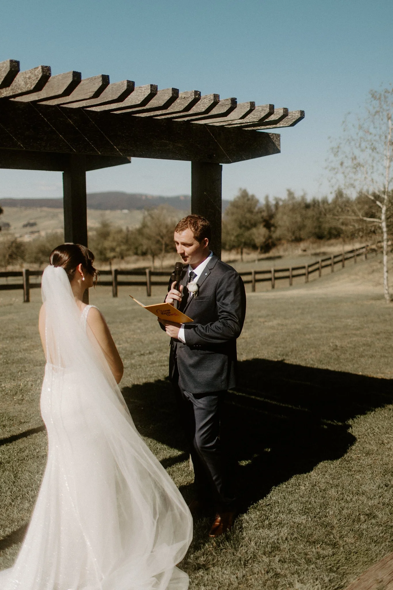 A wedding ceremony taking place outdoors with a bride and groom under a wooden arbor, in a scenic field with trees and mountains in the background at Seclusions Blue Mountains.