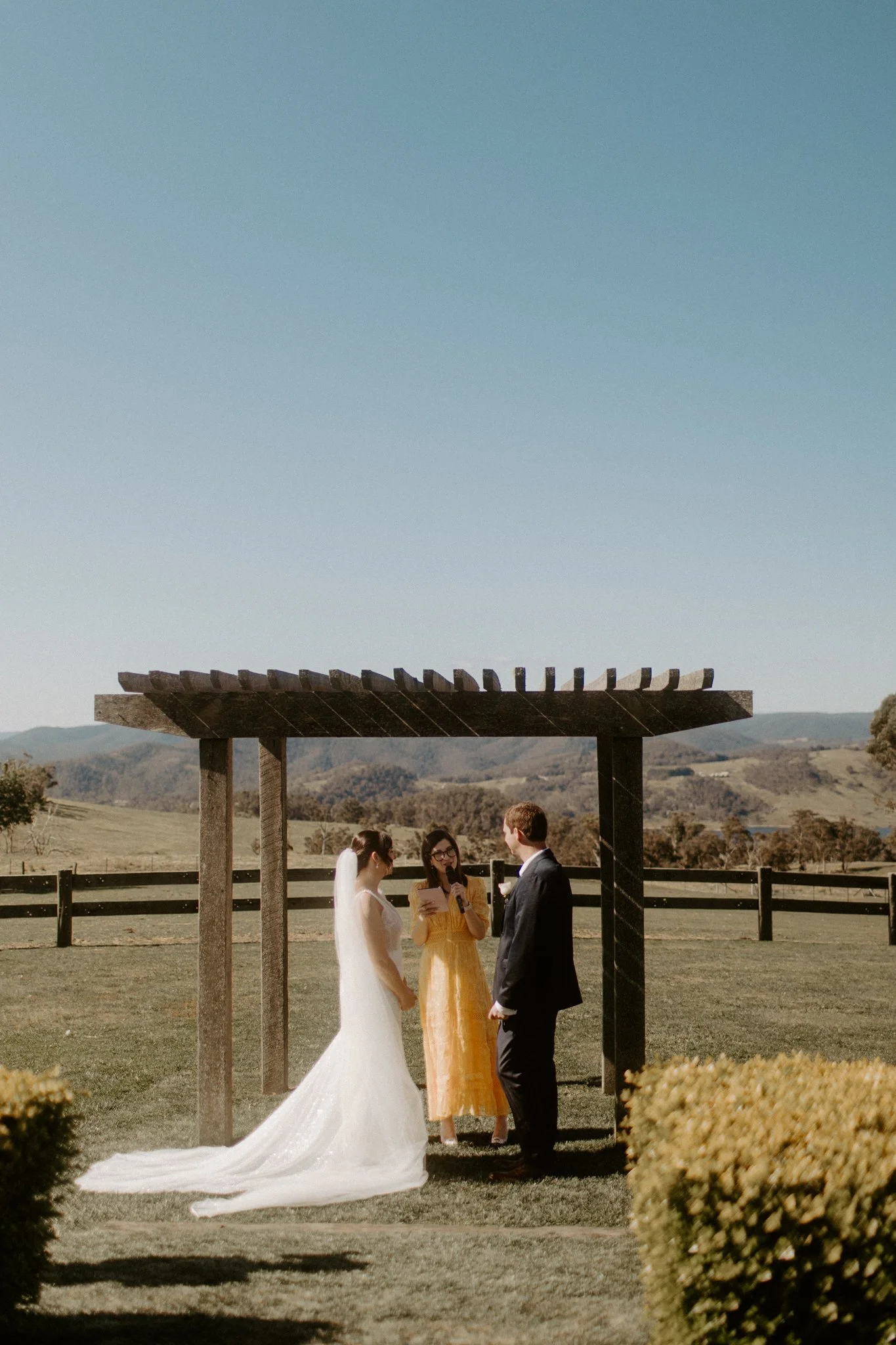 A wedding ceremony taking place outdoors with a bride and groom under a wooden arbor, in a scenic field with trees and mountains in the background at Seclusions Blue Mountains.